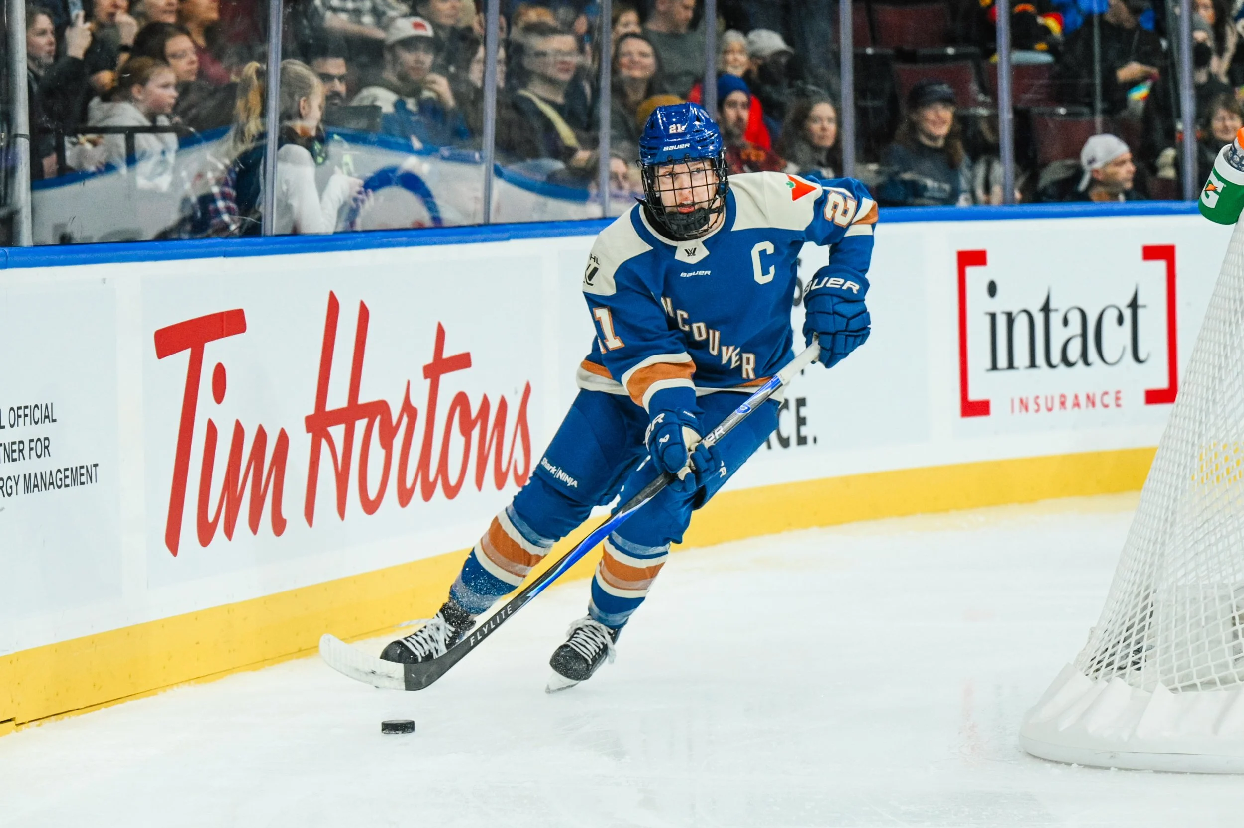 An ice hockey player wearing a blue and white uniform with a captain's 'C' on his chest, skating on the ice near the goal, with spectators watching from the stands in the background.