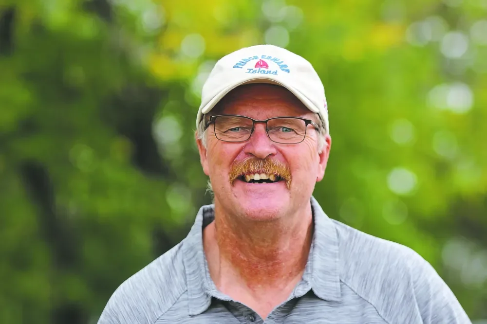 A smiling middle-aged man with glasses and a mustache wearing a baseball cap and a gray collared shirt outdoors with green trees in the background.