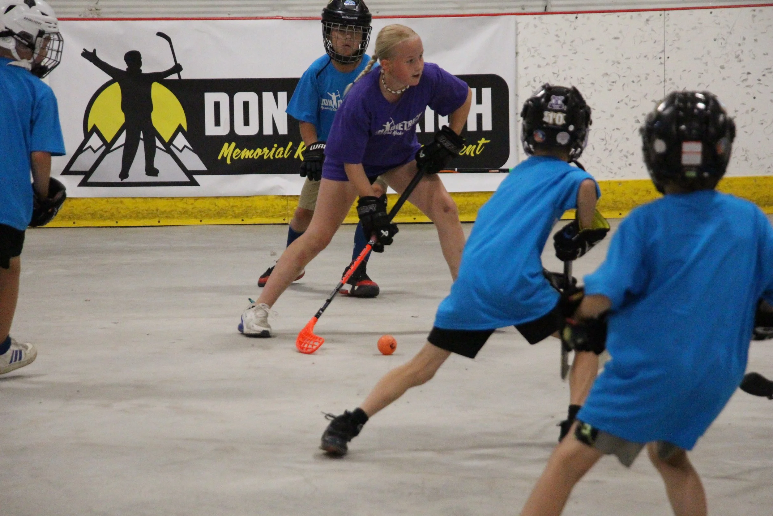 Young girls playing hockey on an indoor rink, with all players wearing helmets and hockey gear, focused on the puck on the ice.