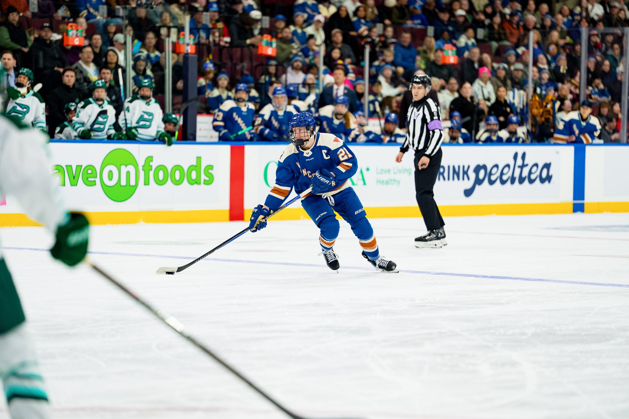 A hockey game in progress with a player in blue jersey number 21 and a captain's 'C' on the front, skating with the puck near the boards, while a referee observes nearby. The background shows spectators sitting in stands, and the rinkside advertising displays.