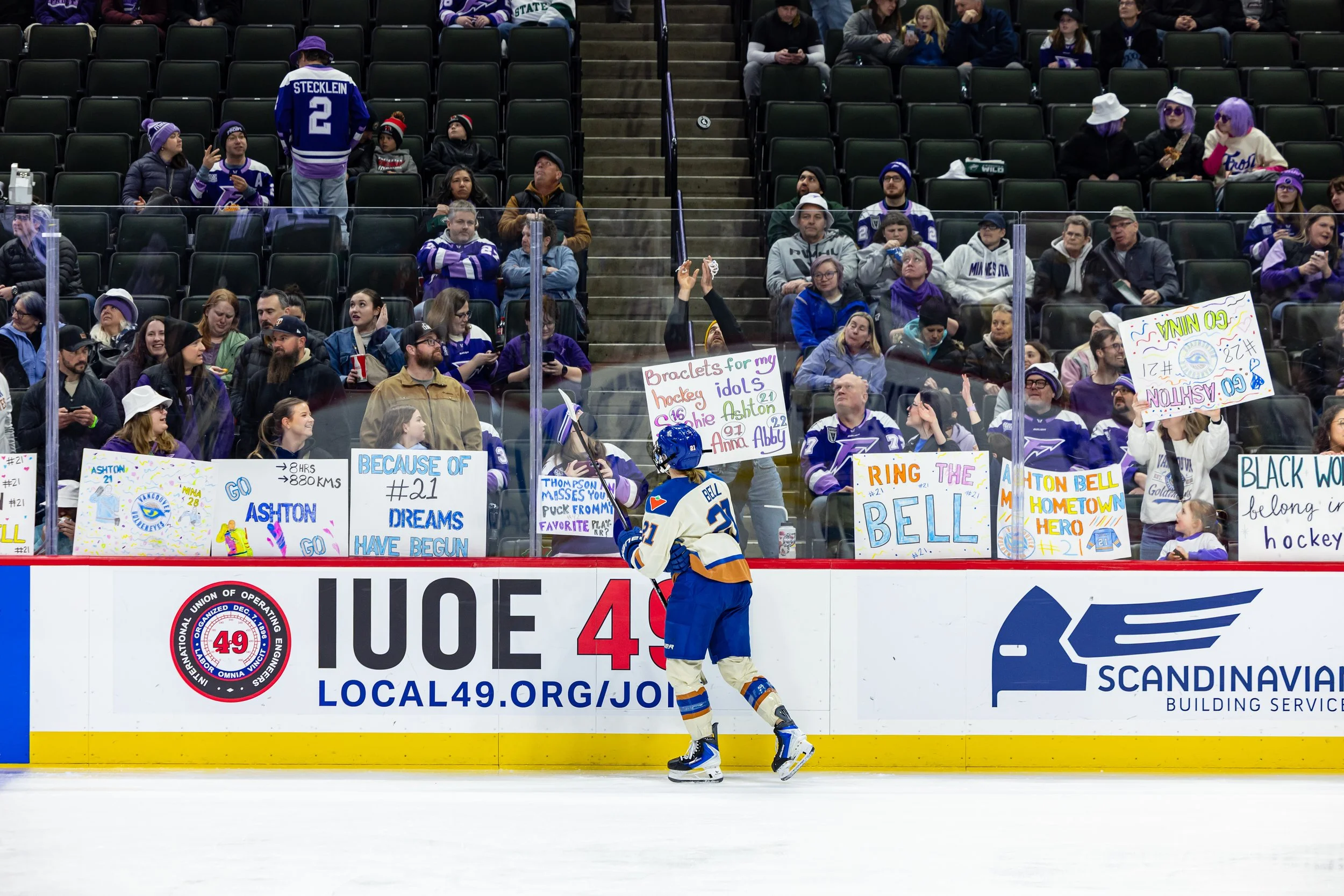 An ice hockey arena with fans in the stands, many wearing purple and hockey jerseys. A hockey player in a blue and yellow uniform is on the ice near the boards, and fans hold colorful signs supporting players and expressing enthusiasm for the game.