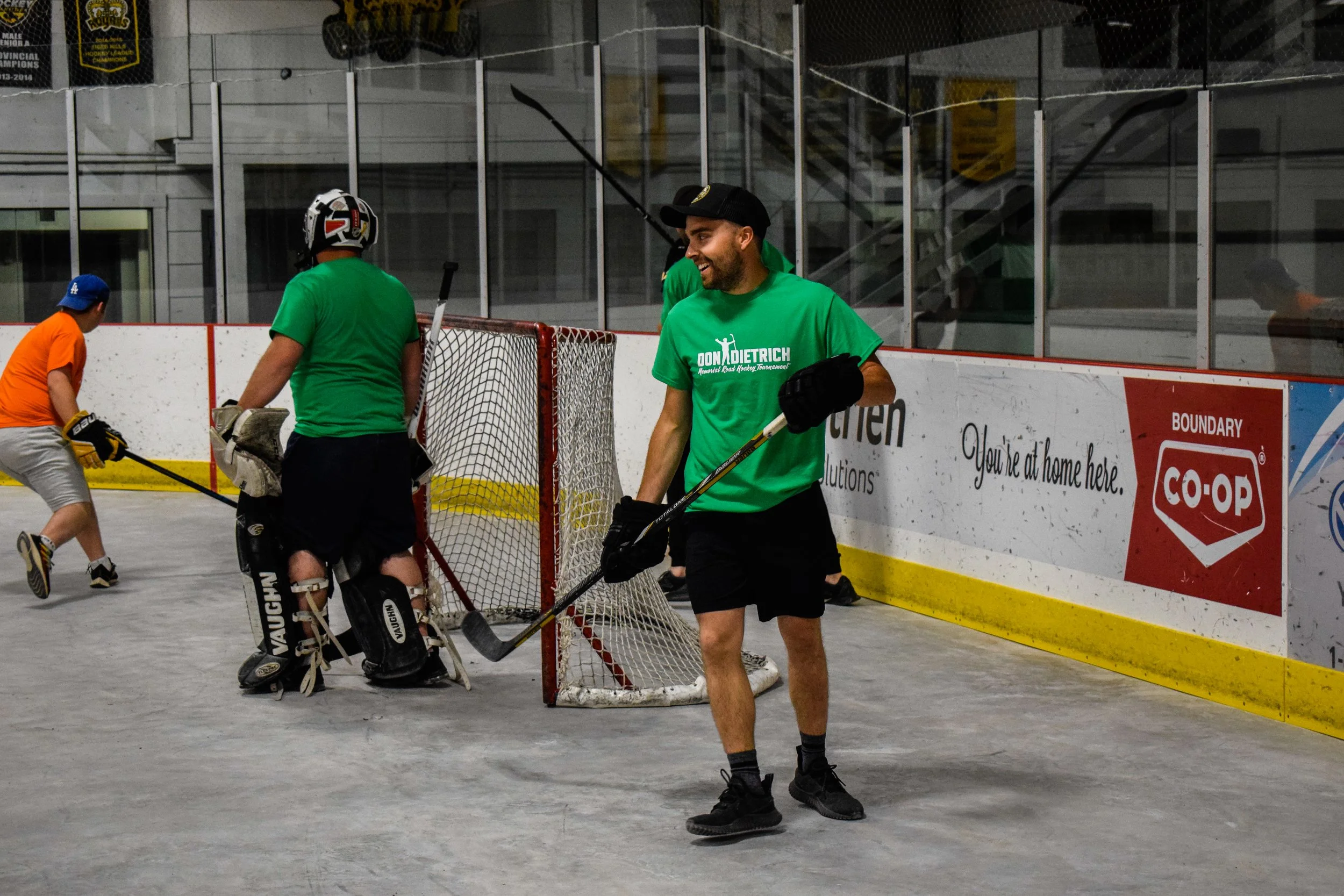 A group of hockey players and a person in casual clothes on an ice rink. The players are wearing colorful shirts and hockey gear, including helmets and gloves. One person in a green shirt and black cap is smiling and holding a hockey stick, standing near the goal net. The background shows the rink's boards, glass, and advertisements.