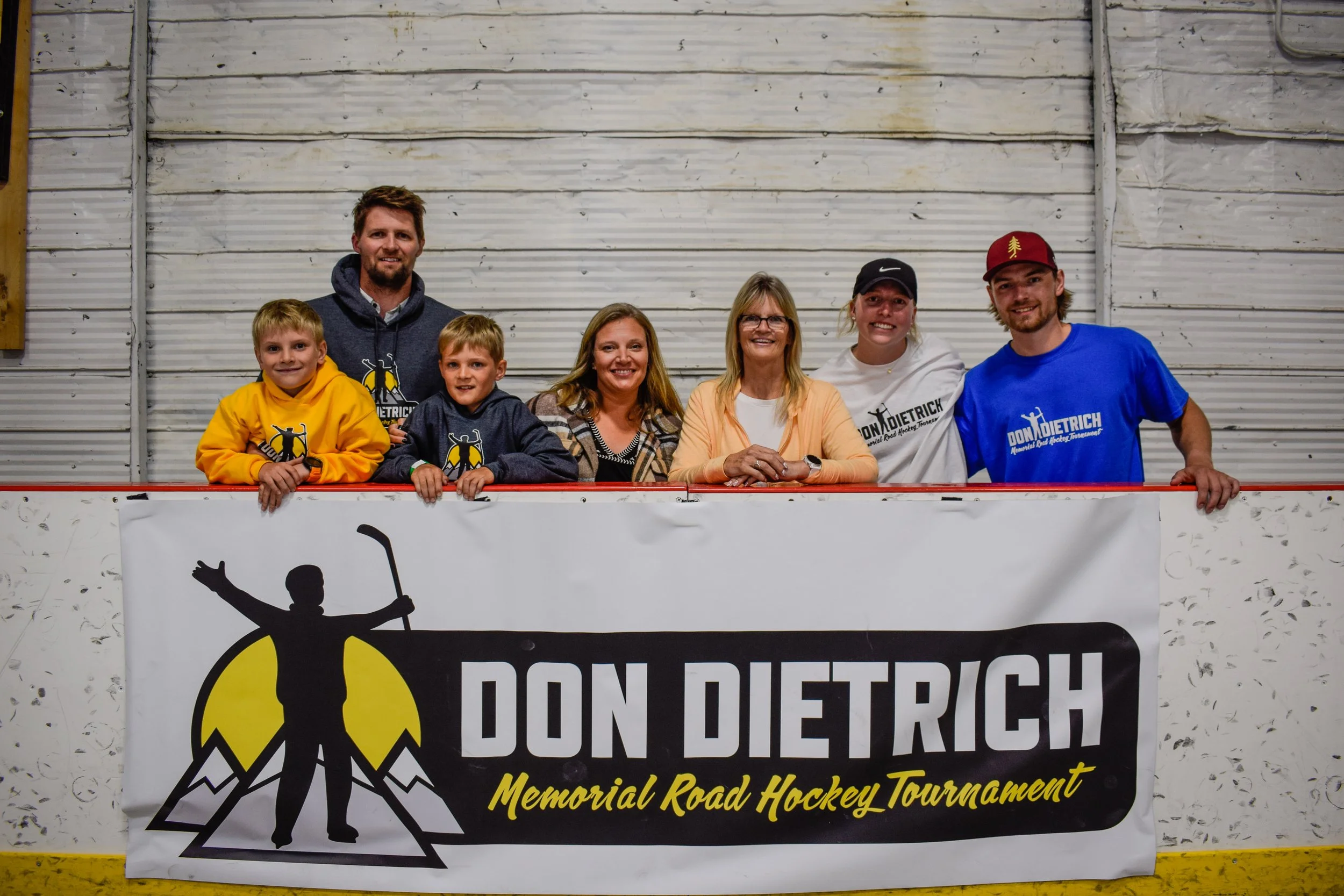 Group of seven people, including children and adults, standing behind a hockey rink barrier at an indoor file with a worn wooden wall background. They are smiling and appear to be participating in a hockey tournament. They are wearing shirts and hoodies with the Don Detrick logo and text for a memorial road hockey tournament.