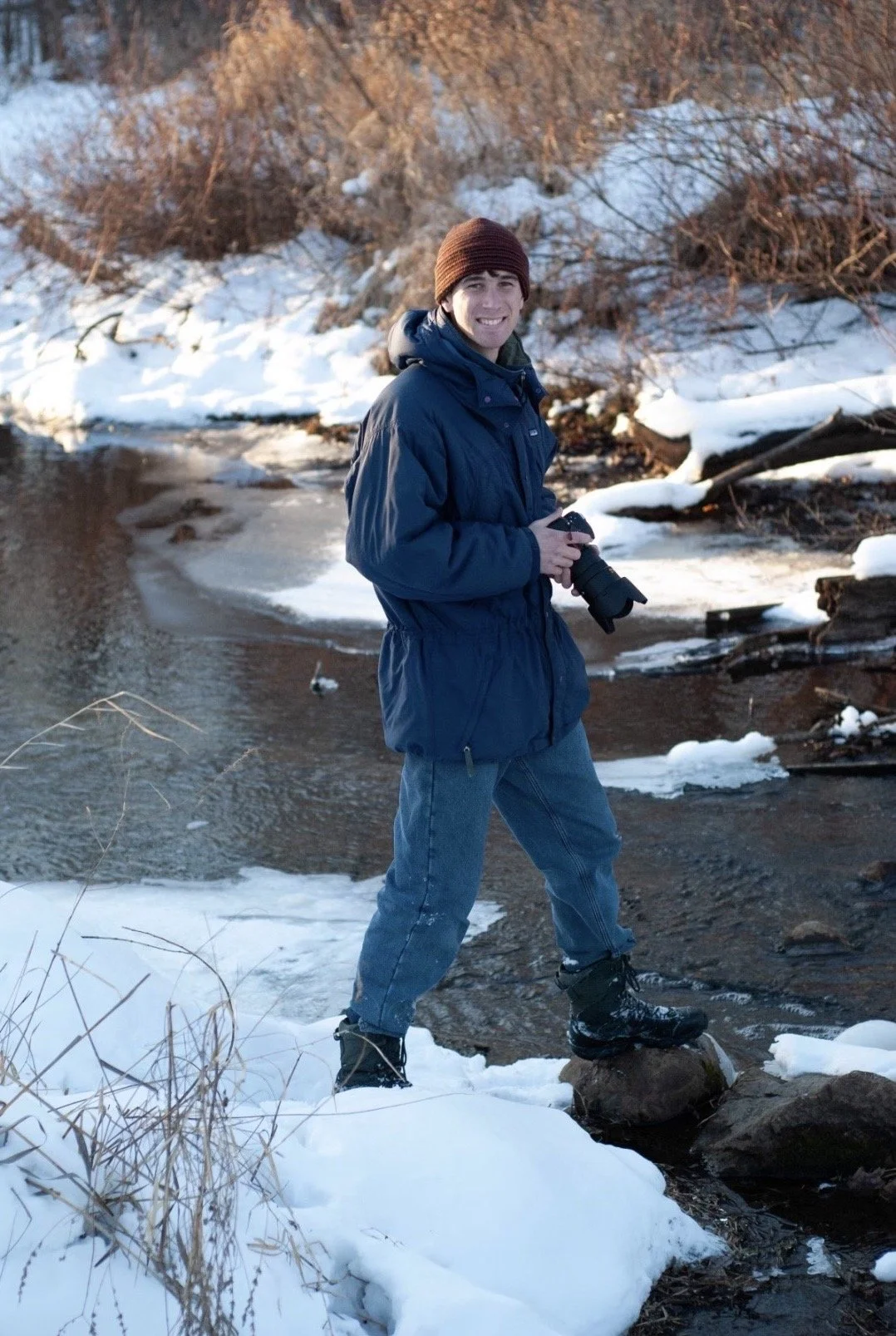 A young man standing in a snowy landscape by a small river, holding a camera, wearing a brown beanie, blue jacket, jeans, and winter boots, smiling at the camera.