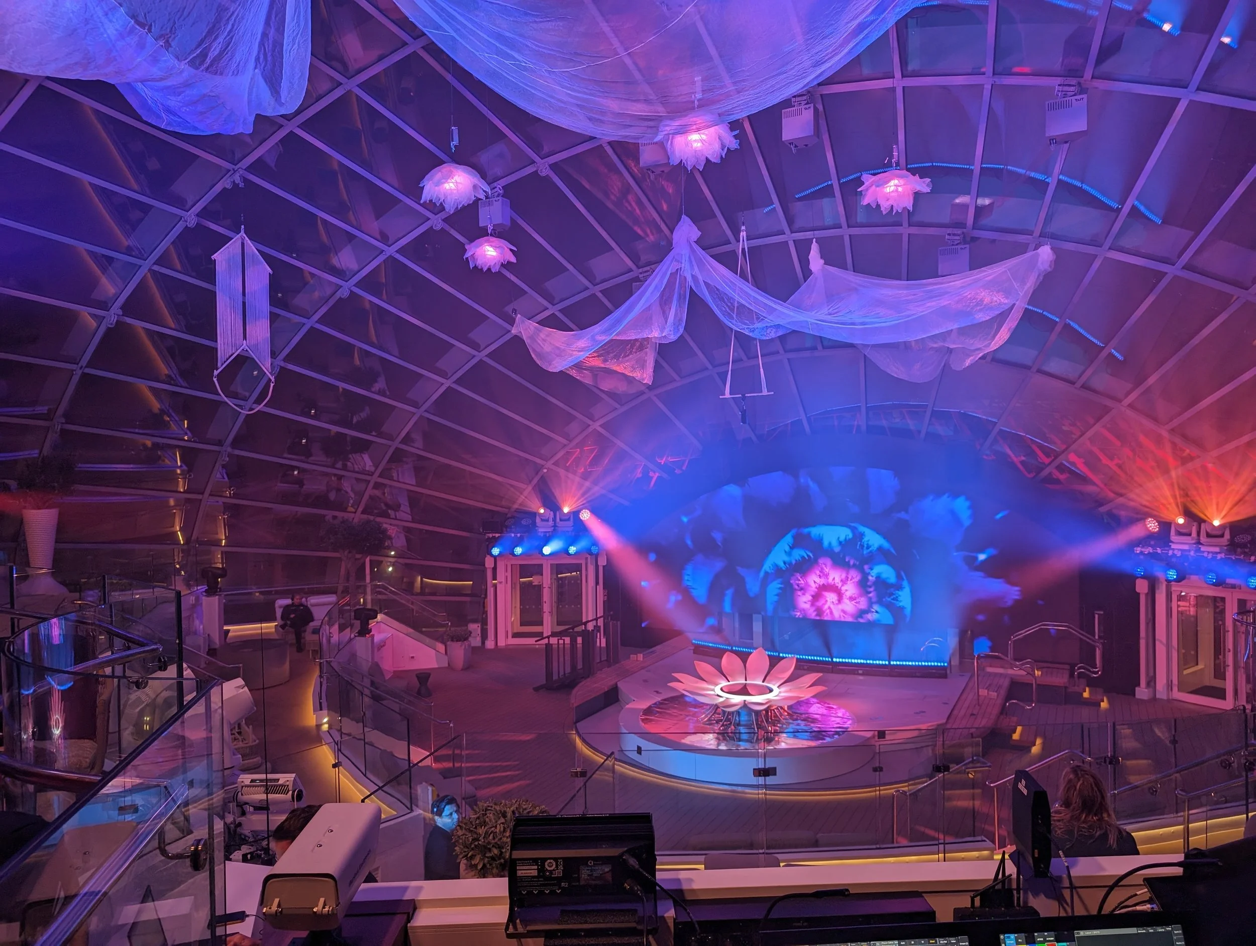 Interior of a building with a glass ceiling, decorated with hanging fabric and flower-shaped lights in purple and pink hues, with a stage and colorful lights.