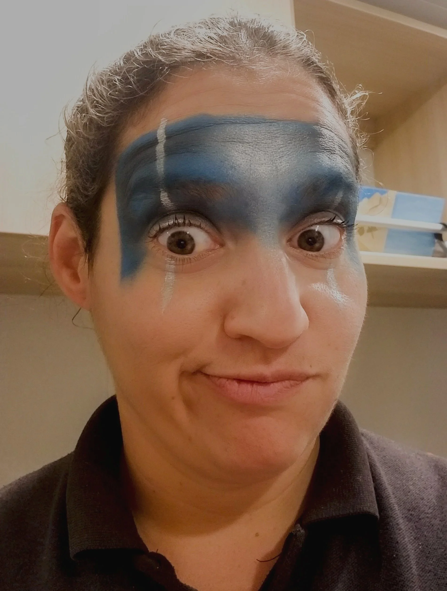 Close-up of a person's face with blue and white face paint around their eyes, wearing a dark top and standing indoors.