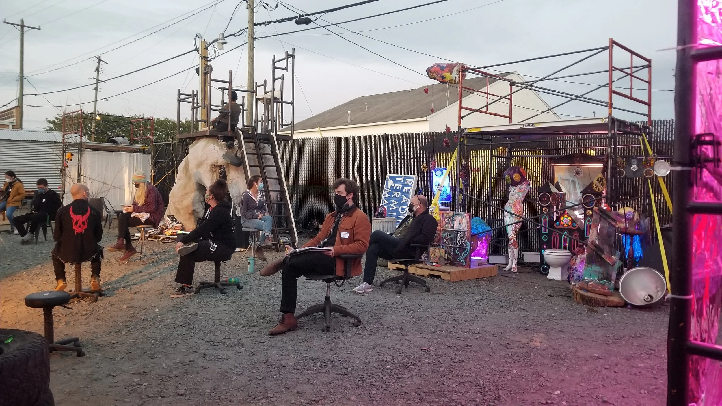 Outdoor art event with people sitting on chairs, some wearing masks, and various artwork and sculptures displayed, including mannequins and electronic decorations, surrounded by metal scaffolding and power lines.