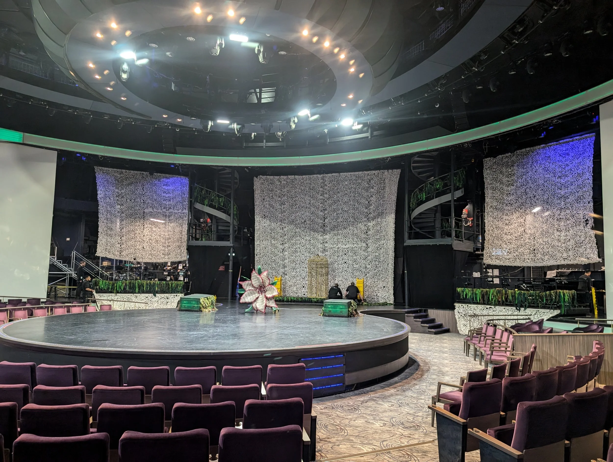 An empty stage in a large auditorium with purple chairs arranged in front. The stage has a circular platform and a floral decoration in the center. The background features textured wall panels and greenery, with equipment and staff setting up behind the scenes.