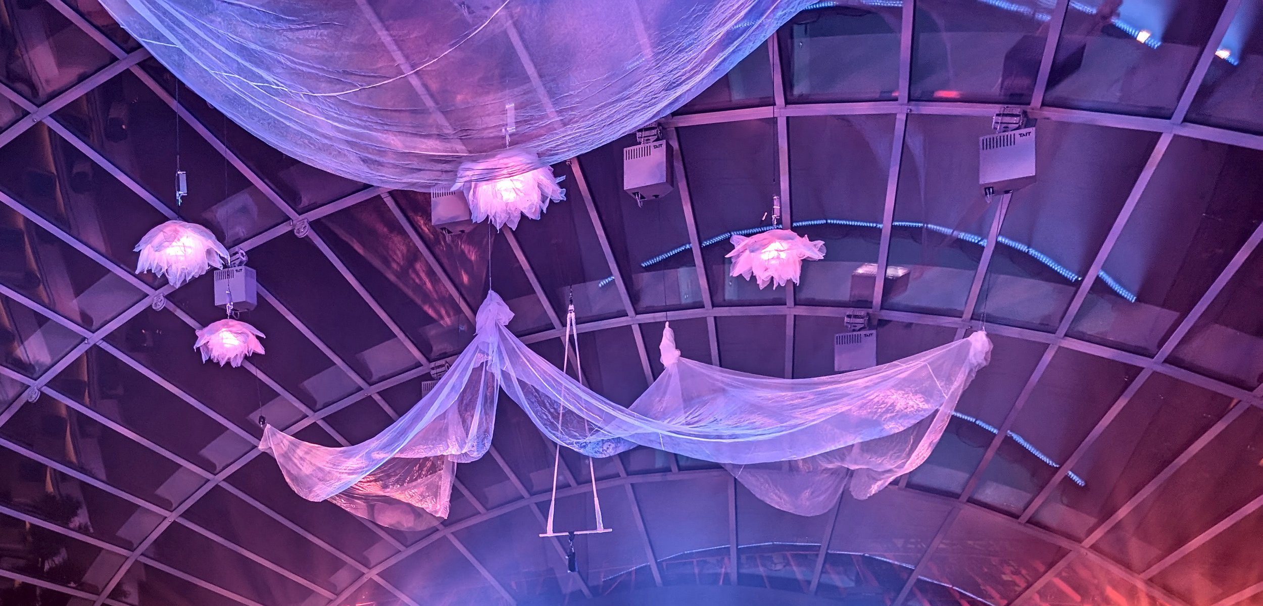 Purple and pink illuminated fabric and flower-shaped hanging lights across a curved ceiling with a grid structure.