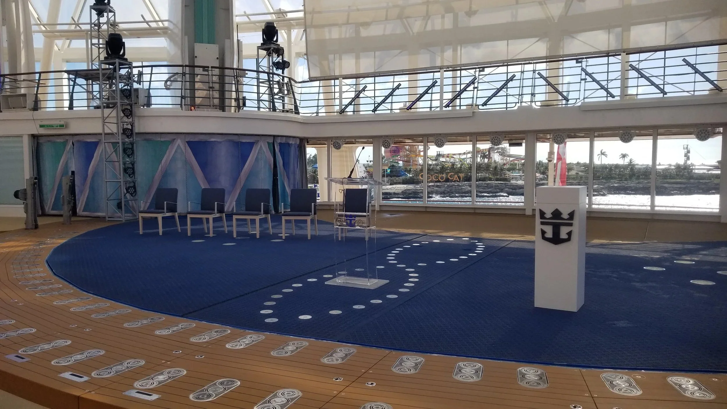 Interior of a boat or cruise ship with a stage and chairs, large windows showing outdoor water and palm trees, and lighting and sound equipment.