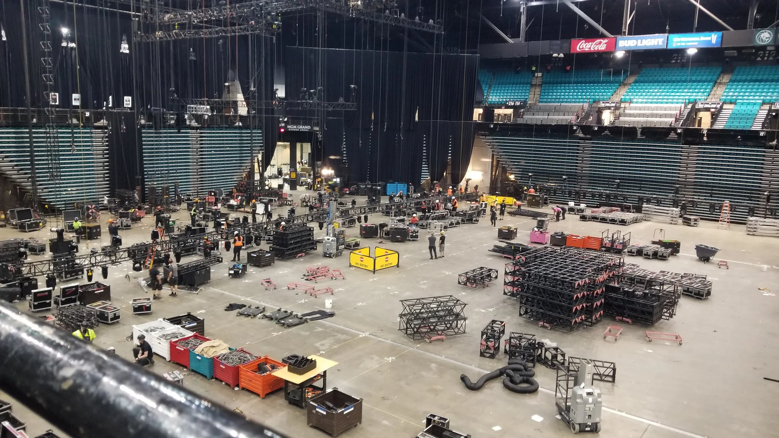 View of an indoor event space with stage setup and numerous equipment cases, scaffolding, and workers preparing for a concert or event at MGM Grand arena.