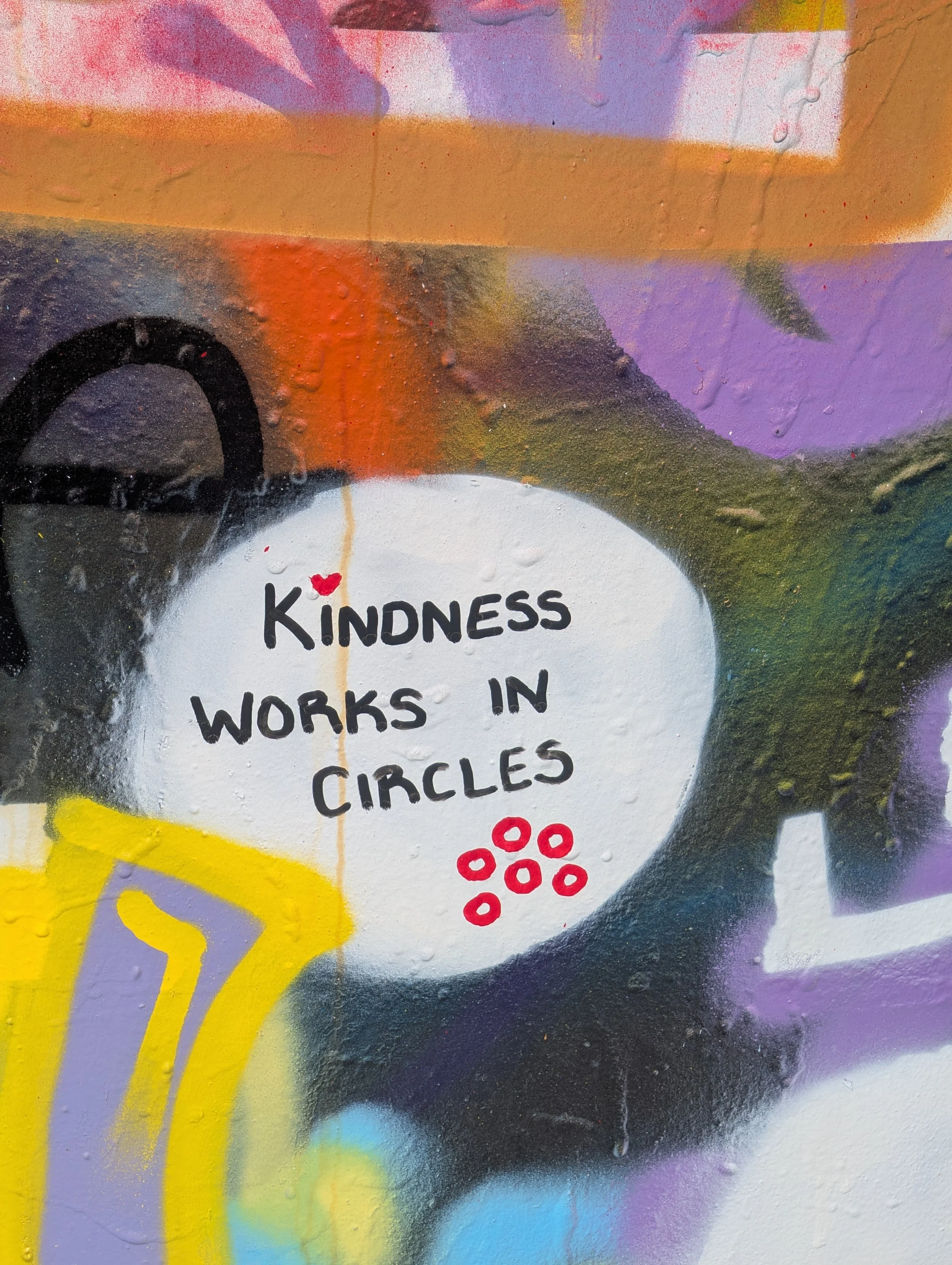 Close-up of a colorful graffiti mural with the phrase 'Kindness Works in Circles' written inside a white circle, surrounded by abstract paint splatters and geometric shapes in purple, yellow, black, red, and orange.