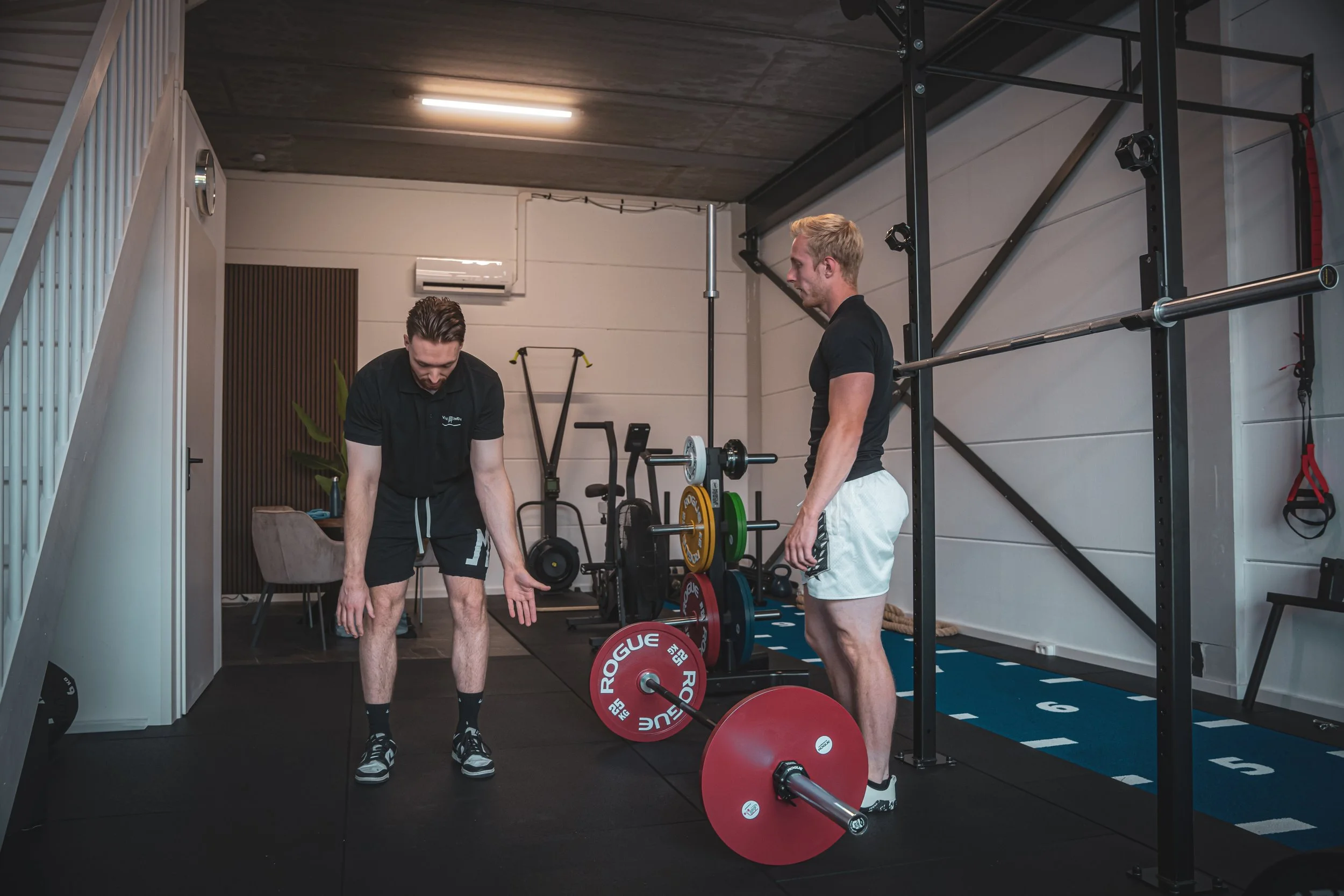 Twee mannen in een fitnessruimte, de ene is aan het bukken en de andere kijkt toe, omringd door fitnessapparatuur en gewichten.