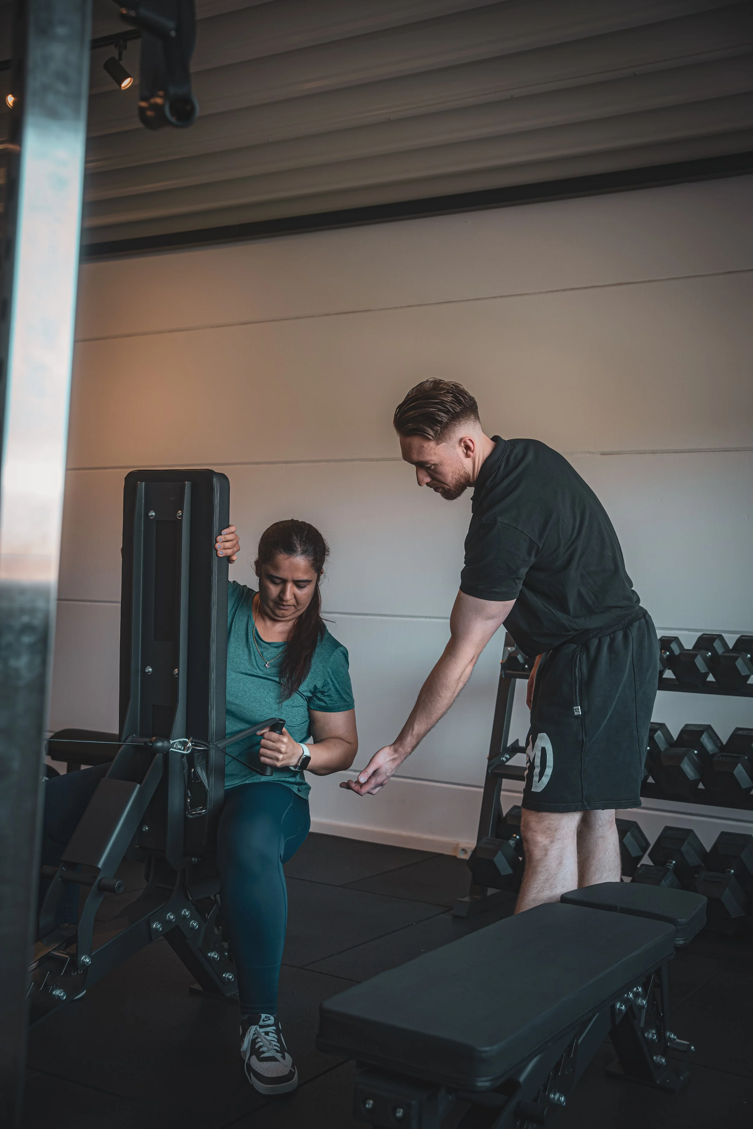 Een man en een vrouw in een sportschool, de vrouw zit op een apparaat en kijkt op haar telefoon terwijl de man naast haar zich naar haar buigt en met haar praat.