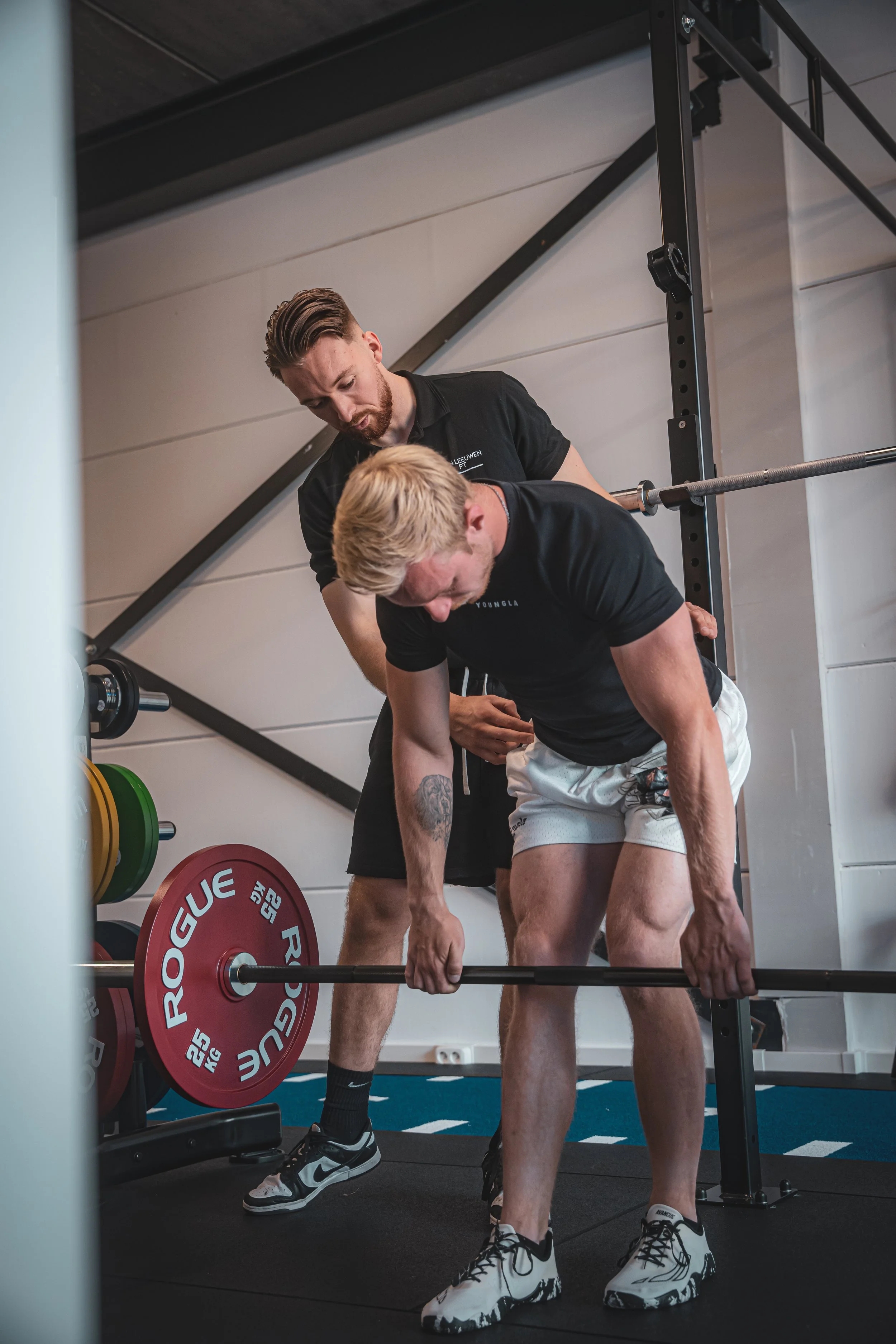 Twee mannen die samen een gewicht heffen in een sportschool, de ene man geeft instructies terwijl hij op een bankje staat en de ander de barbell tilt.