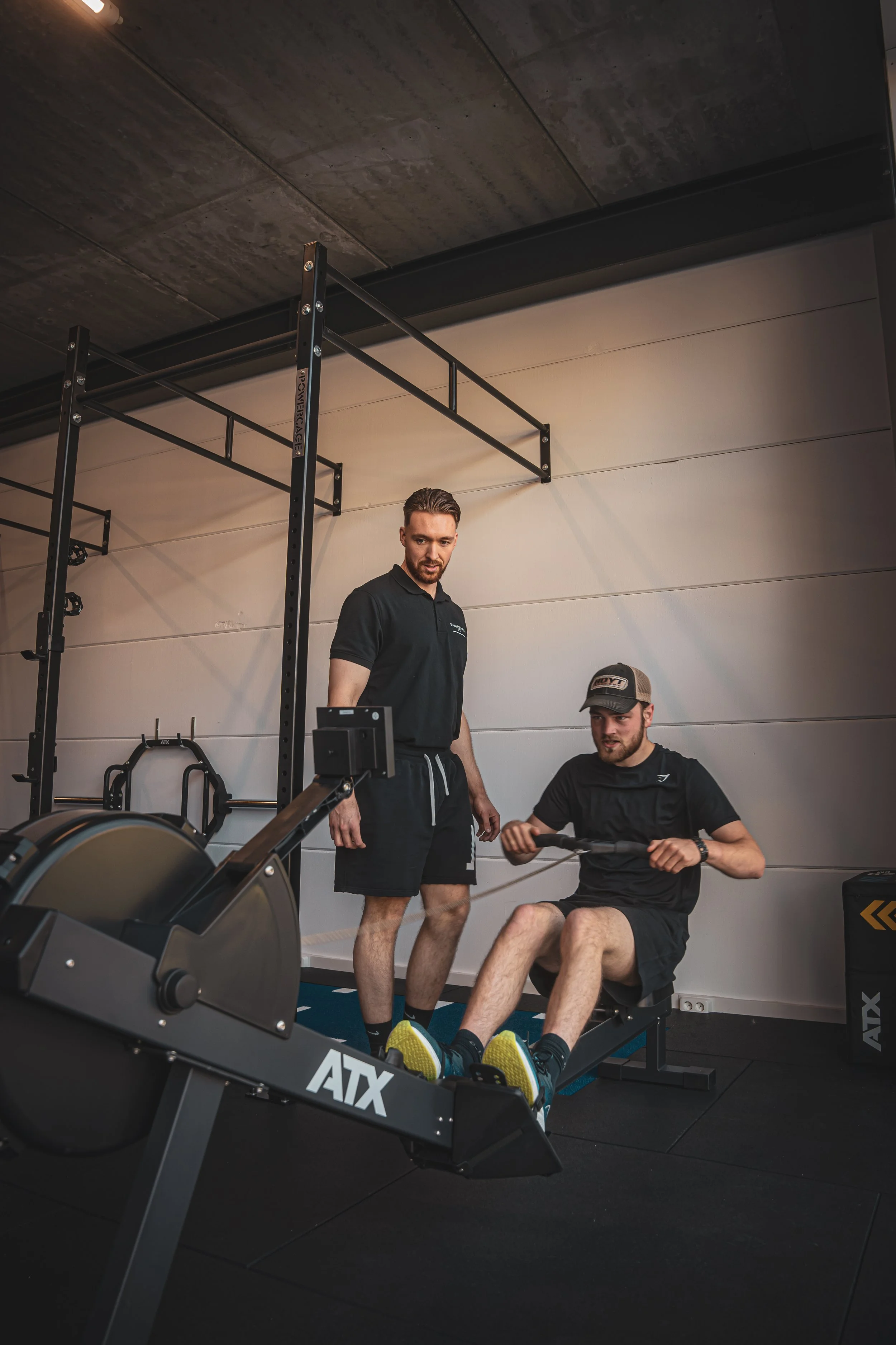 Twee mannen in een sportschool, één persoon zit op een roeitrainer en de andere persoon staat naast hem waarbij hij hem helpt of adviseert.