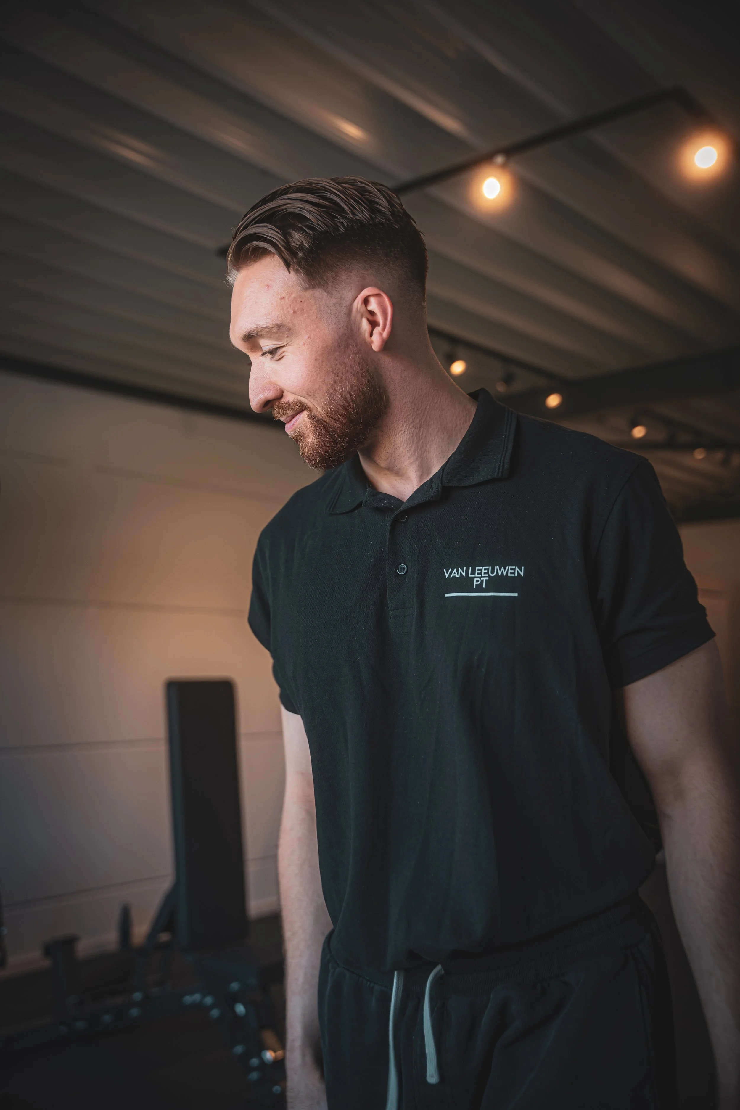 Man met een baard en kort geschoren haar, lachend in een modern interieur met gemakkelijke verlichting, draagt een zwart polo met tekst 'VAN LEEUWEN PT' en doet oefeningen op een fitnessapparaat.