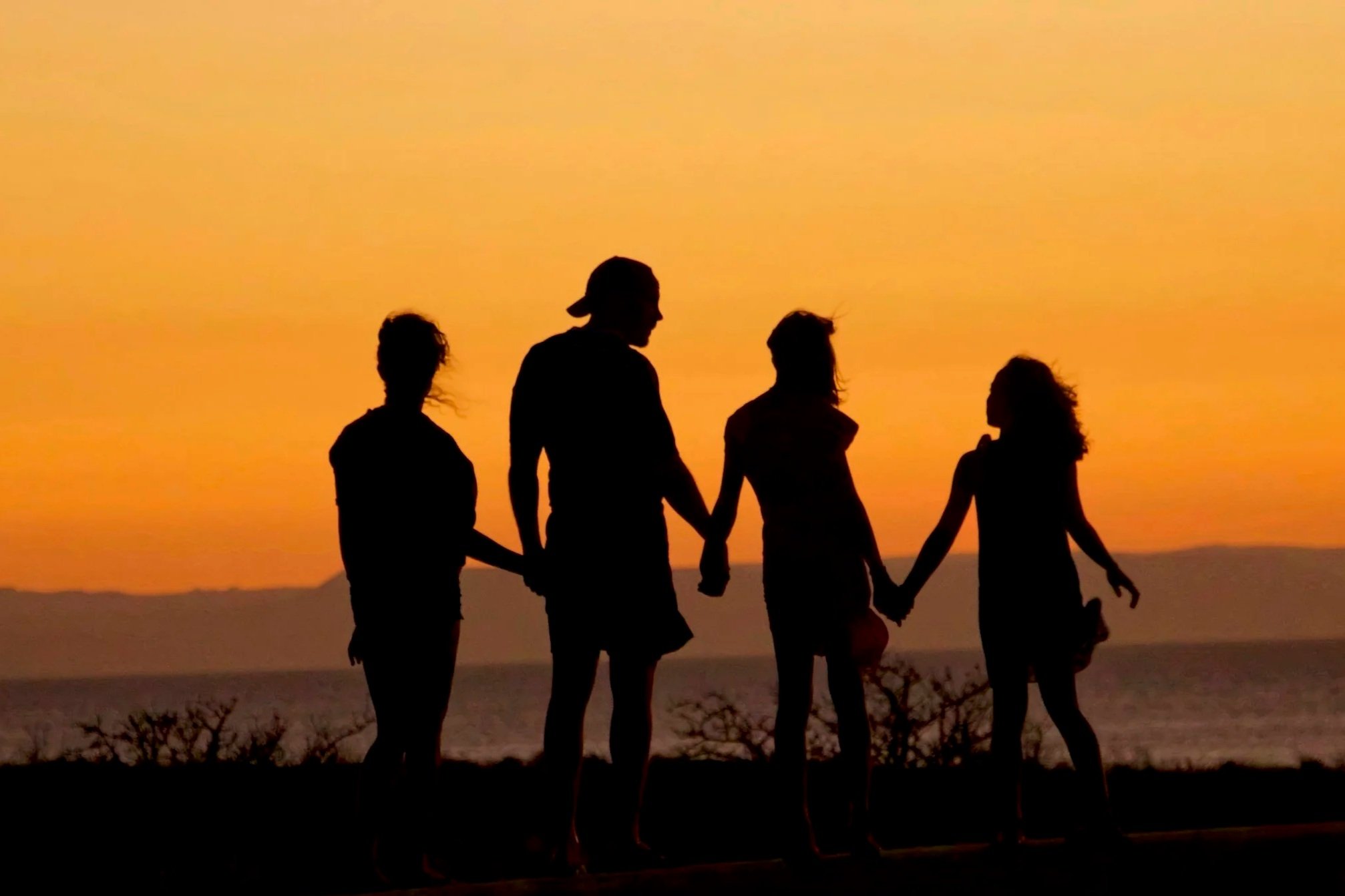 Silhouettes of five people holding hands and walking on the beach at sunset with a vibrant orange sky.