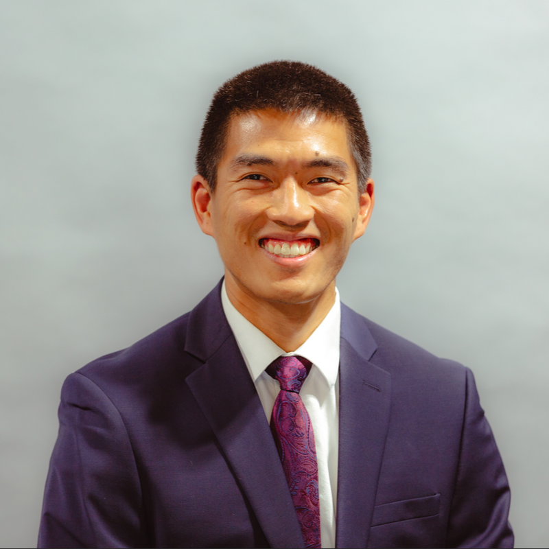 A man in a navy suit and tie smiling in front of a plain background.