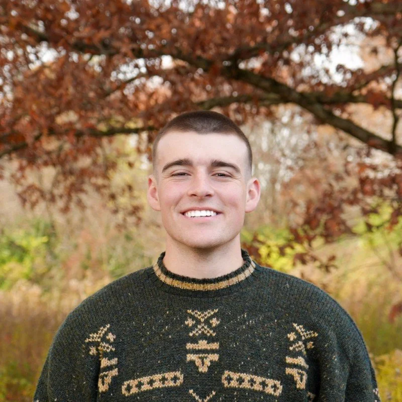 A young man with short hair and a big smile standing outdoors in front of trees with autumn-colored leaves, wearing a dark sweater with a tan pattern.