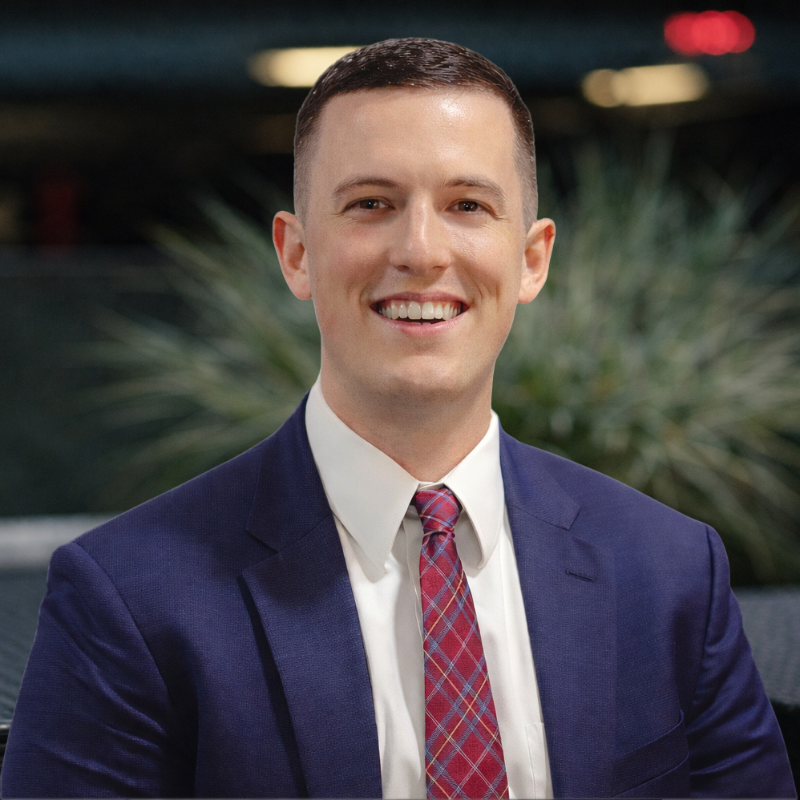 A young man in a navy blue suit, white shirt, and red plaid tie smiling at the camera, with a blurred outdoor background.