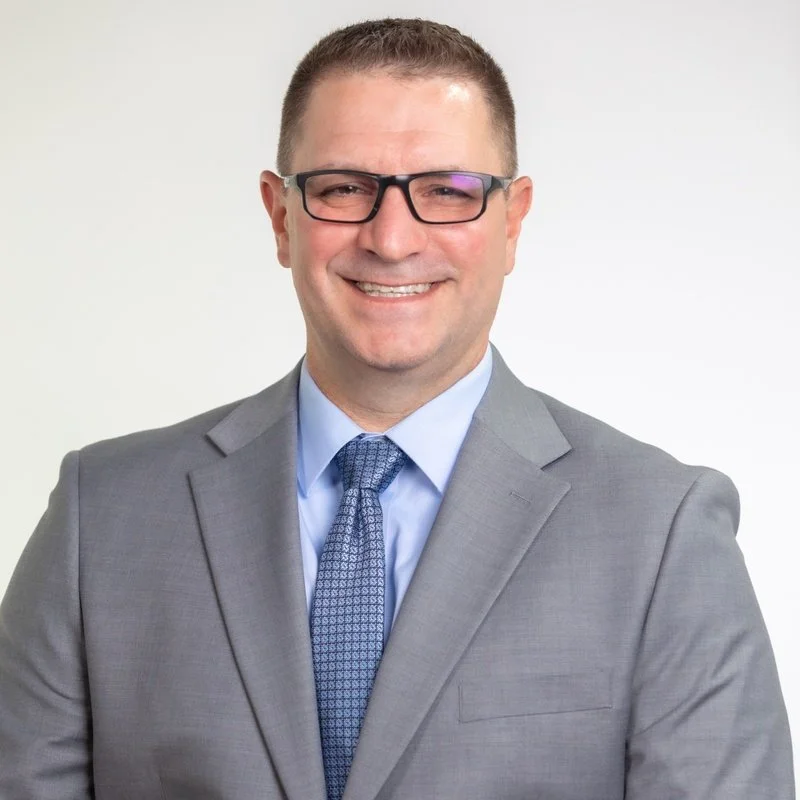Portrait of a smiling man wearing glasses, a gray suit, and a blue tie against a plain white background.