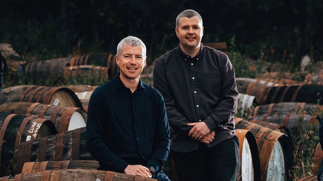 Two men standing among rows of wine barrels outdoors during the daytime, smiling at the camera.