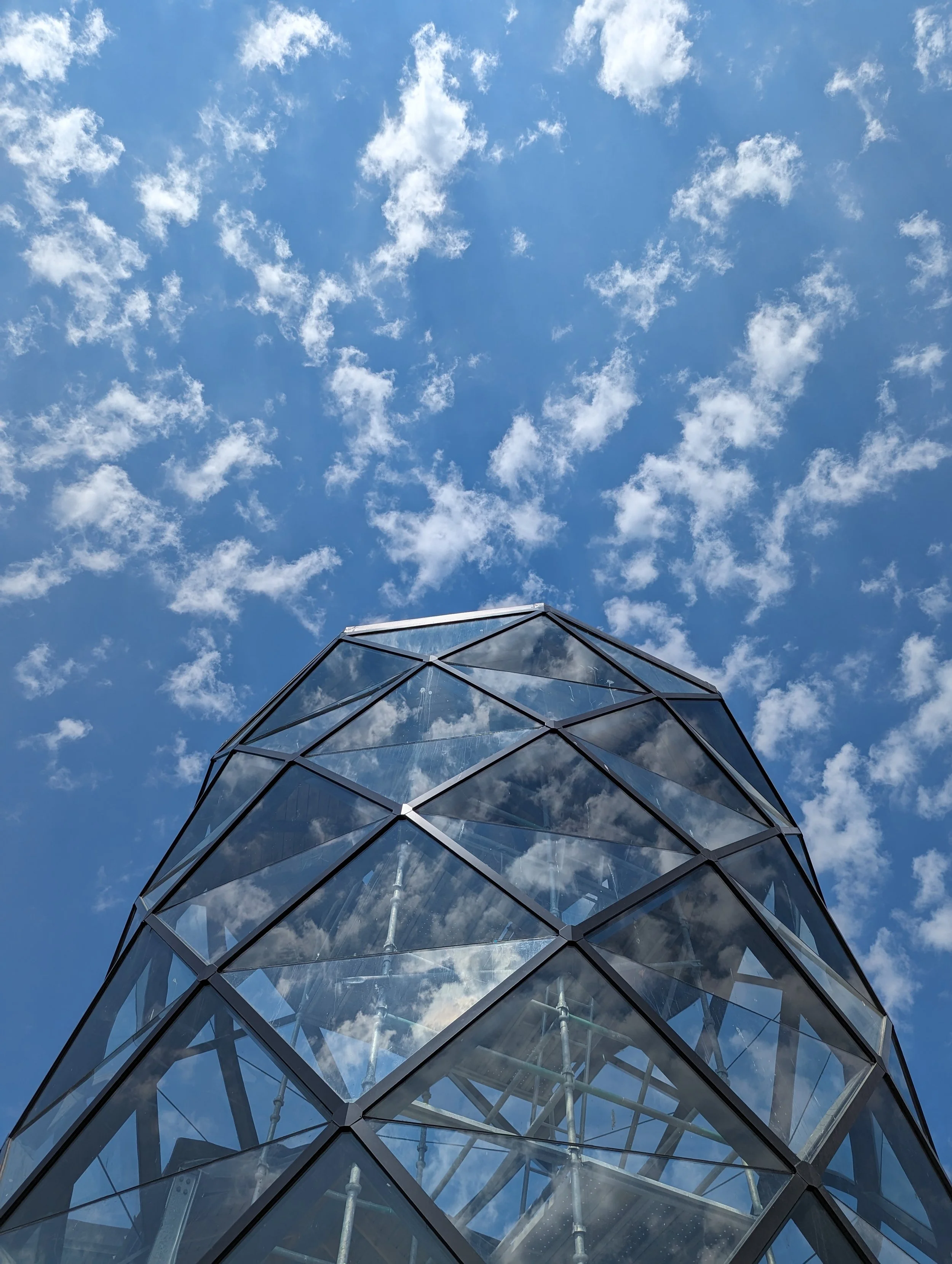 A modern glass building with geometric facades reaching toward a bright blue sky filled with scattered white clouds.
