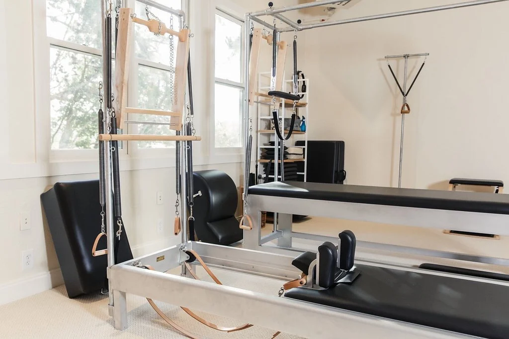 Home Pilates or physical therapy room with a reformer, trapeze, and chairs near windows.