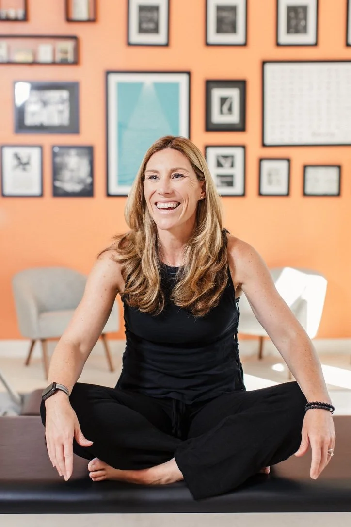 A woman sitting cross-legged on a yoga mat, smiling, in a room with framed artwork on the orange wall behind her.