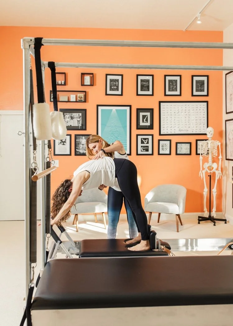 Two women practicing Pilates in a studio, one assisting the other on a reformer machine, with a pastel orange wall decorated with framed artwork and a skeleton model nearby.