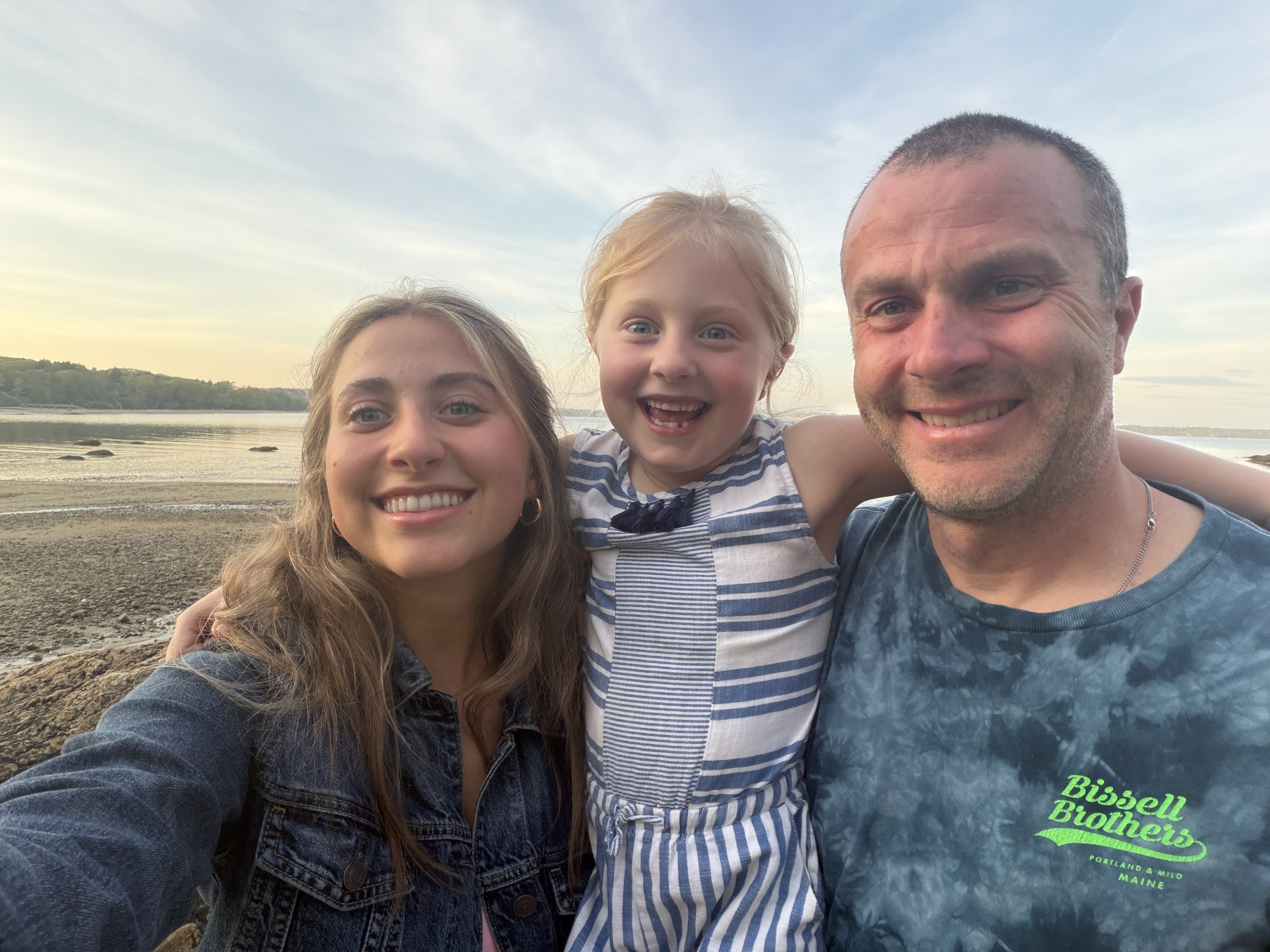 A smiling family of three, including a young girl with red hair, a woman with long brown hair, and a man with short hair, taking a selfie on a beach during sunset. The background features a calm body of water, rocks, and a cloudy sky.