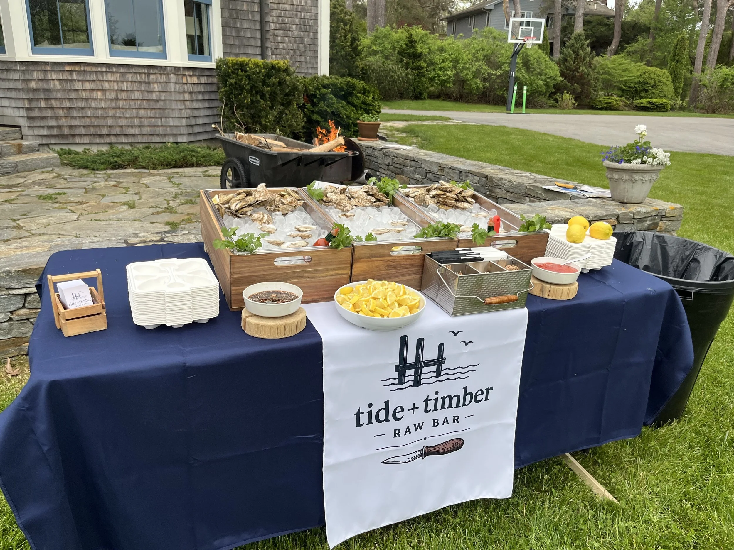 Outdoor bar setup with oysters on ice, lemons, sauces, and utensils, with a fire pit and house in the background.