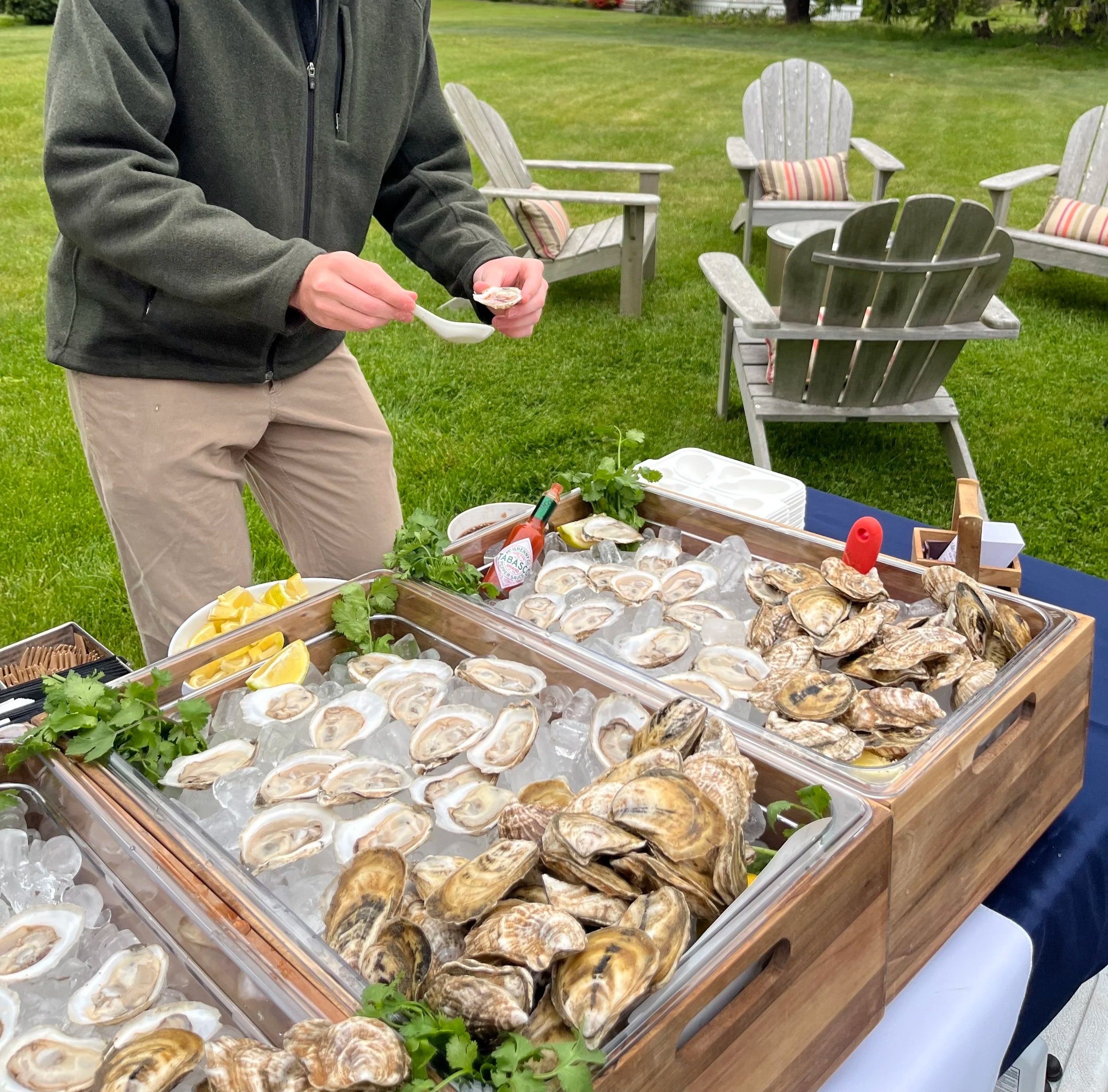 Part of a person in a green jacket and khaki pants serving oysters at an outdoor seafood stand with various lemon slices, hot sauce, and garnishes on the table.