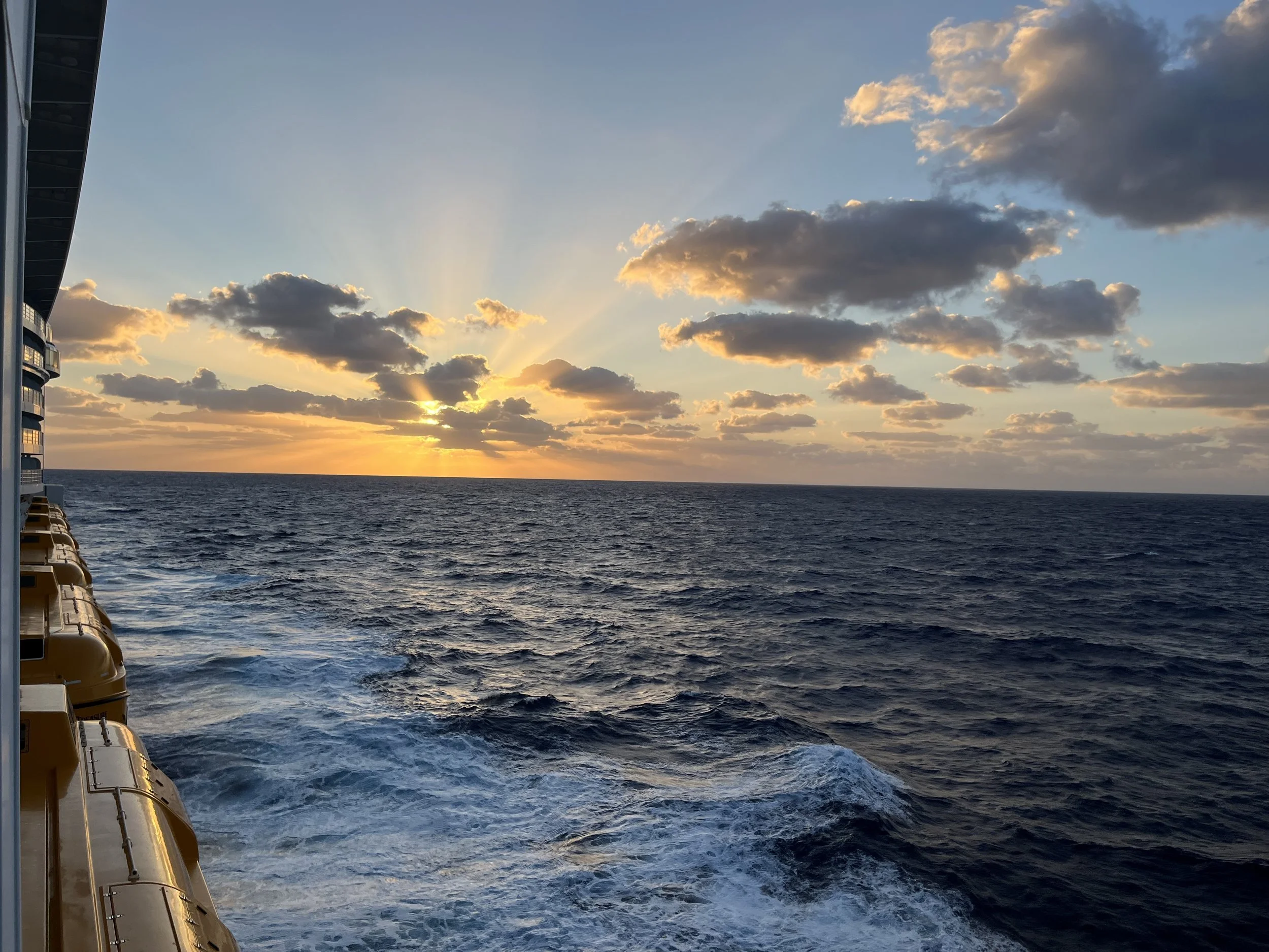 View of the ocean at sunset or sunrise from a cruise ship deck, with clouds in the sky and the ship's side with yellow lifeboats visible on the left.