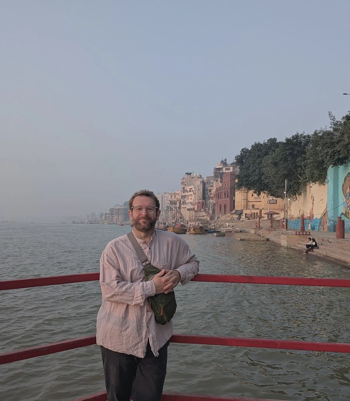 A man standing at a ghat in Varanasi alongside the river Ganges, with colorful buildings and trees in the background, and a sky with light haze.