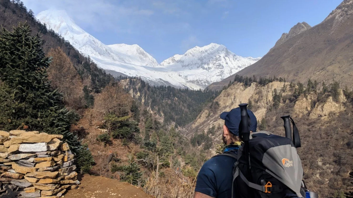Hiker with backpack looking towards snow-capped mountains and forested valley