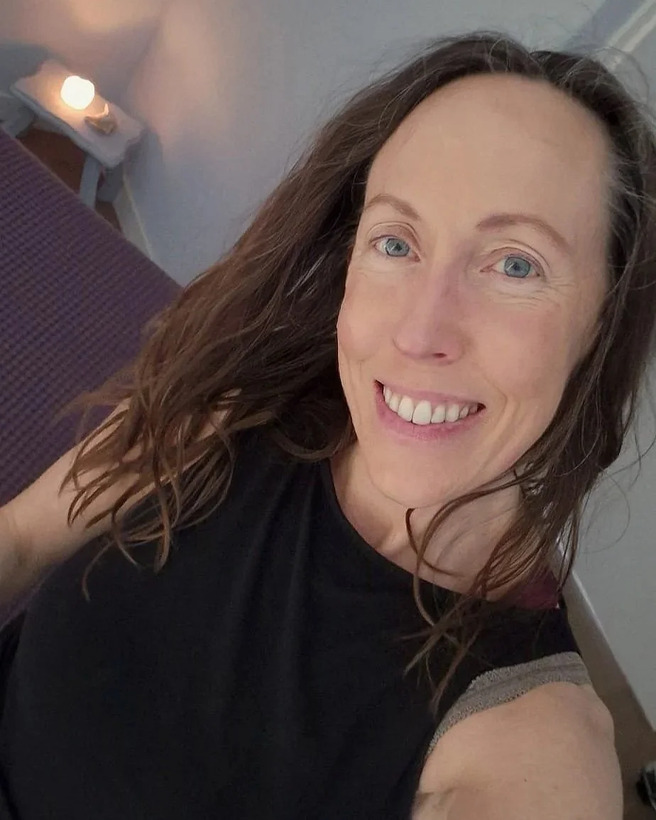 A woman with long, wavy brown hair, blue eyes, and a big smile taking a selfie in a dimly lit room near a bed with a small lamp on a nightstand.