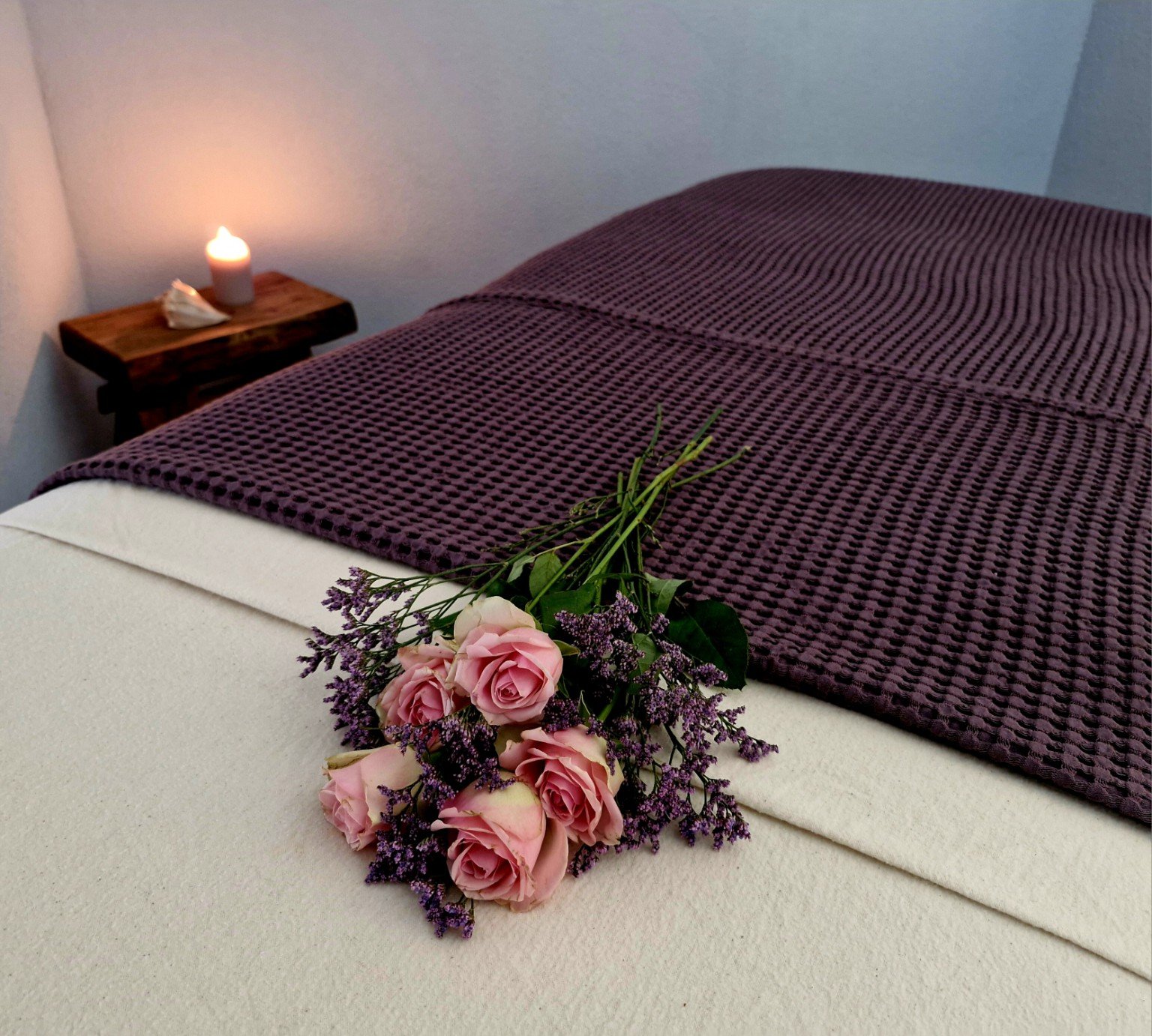 A bouquet of pink roses and purple lavender rests on a bed with a purple textured blanket. In the background, a lit candle and a seashell are on a wooden side table in a cozy, softly lit room.