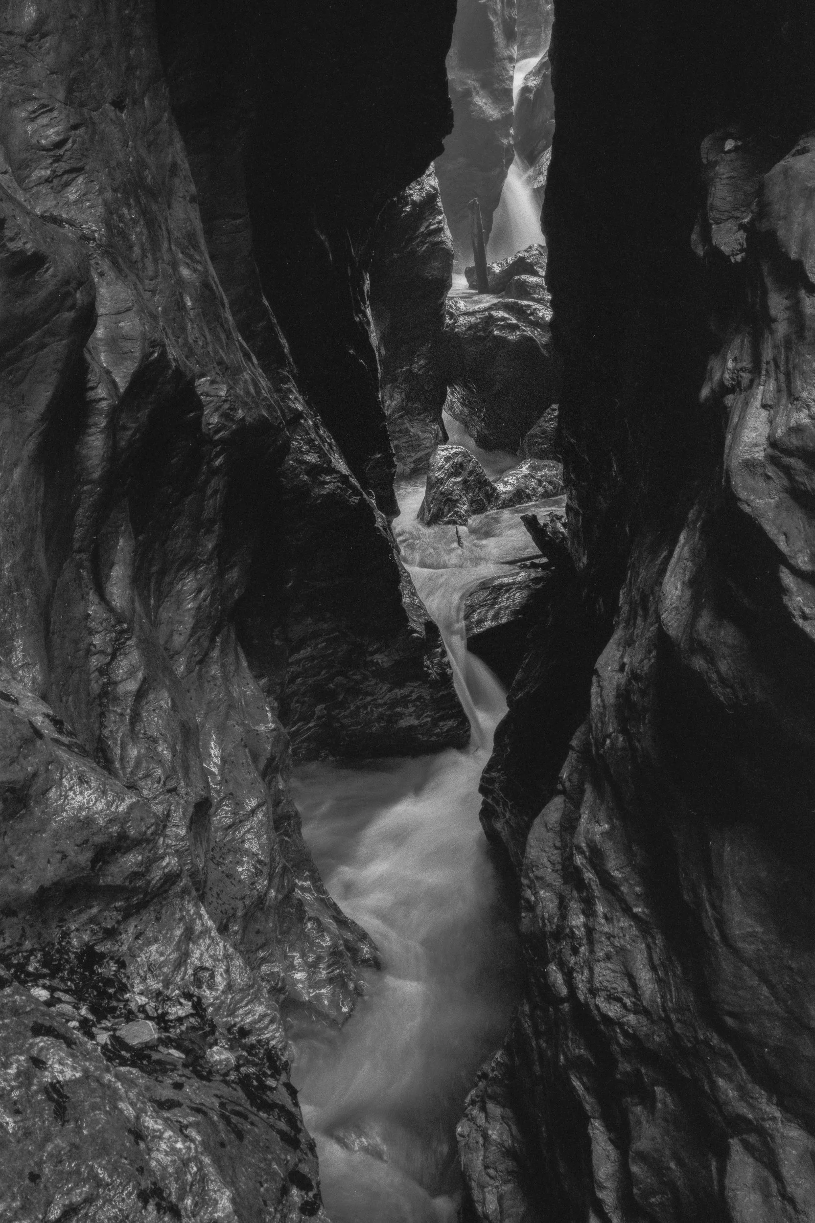 Ein dunkler Canyon mit Wasserfluss zwischen glatten, nassen Felswänden. In der Ferne ist ein Wasserfall sichtbar, der in den Canyon stürzt.
