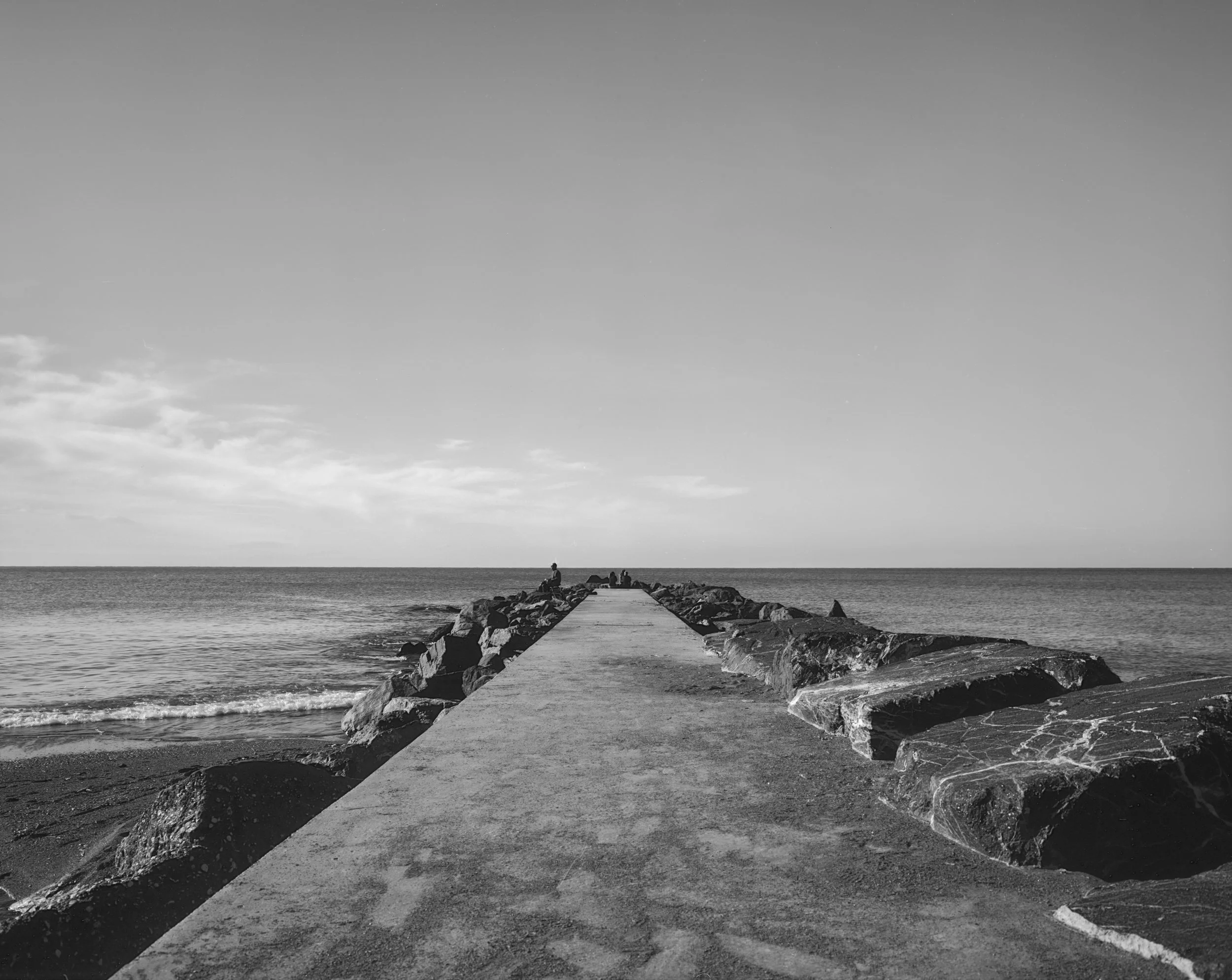 Schmale Mole am Meer mit einer Person, die dort sitzt, im Hintergrund das Wasser und der Himmel mit wenigen Wolken.