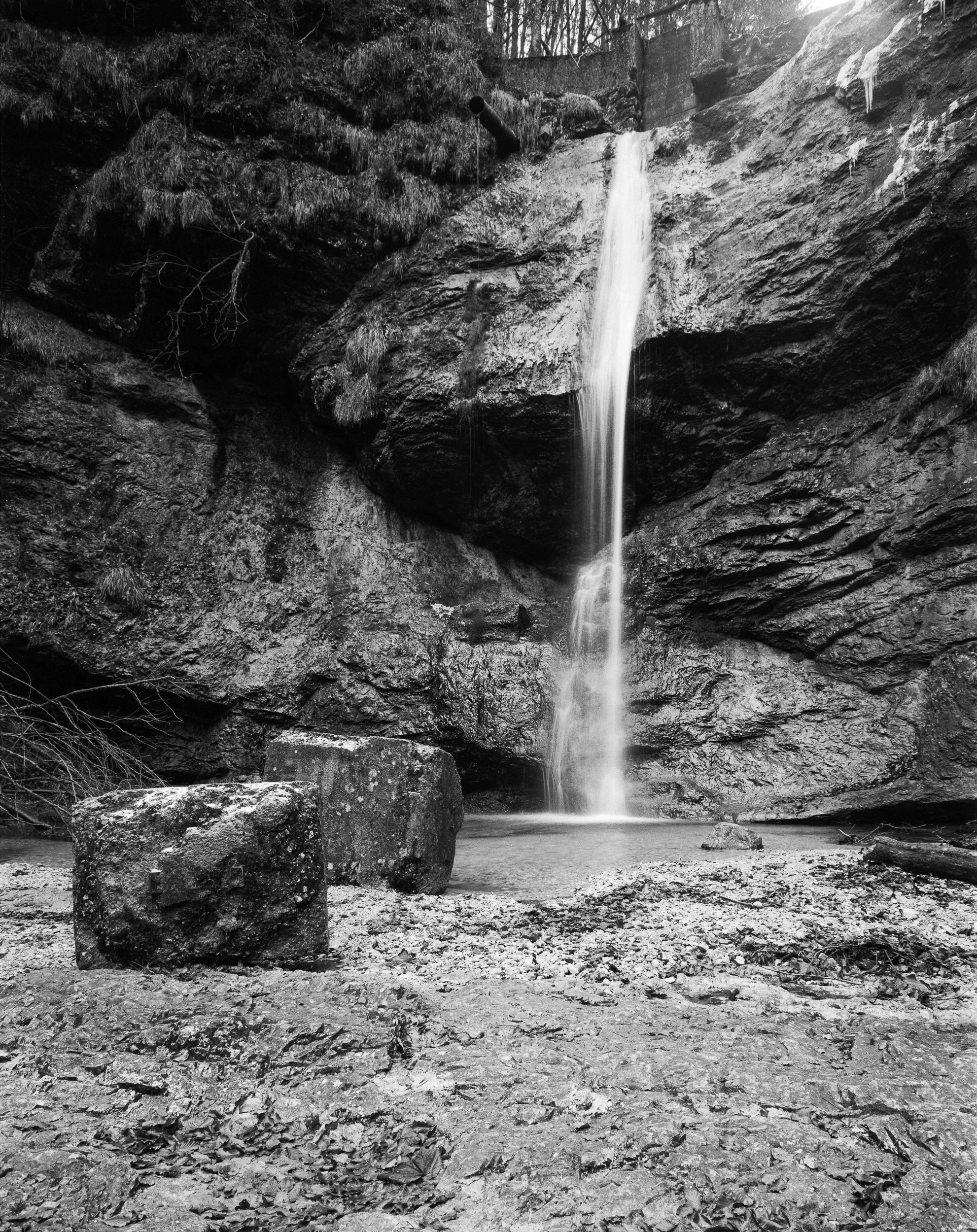 Ein Wasserfall in einer felsigen Landschaft, umgeben von Bäumen und Steinen, in Schwarzweiß fotografiert.