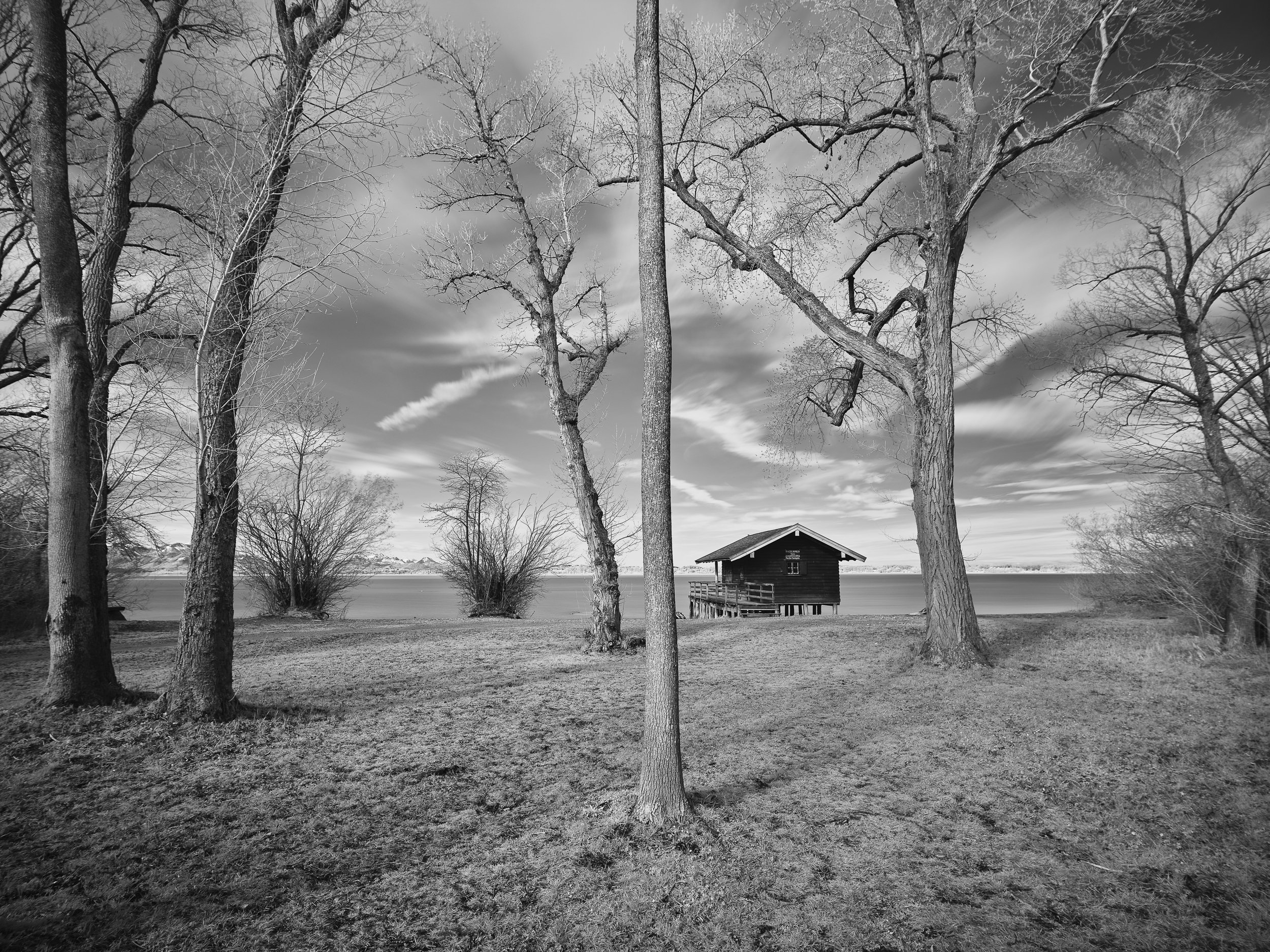 Schwarz-weiß-Bild eines Sees mit Bäumen am Ufer, einem kleinen Holzhaus auf Stelzen im Wasser und einem bewölkten Himmel.