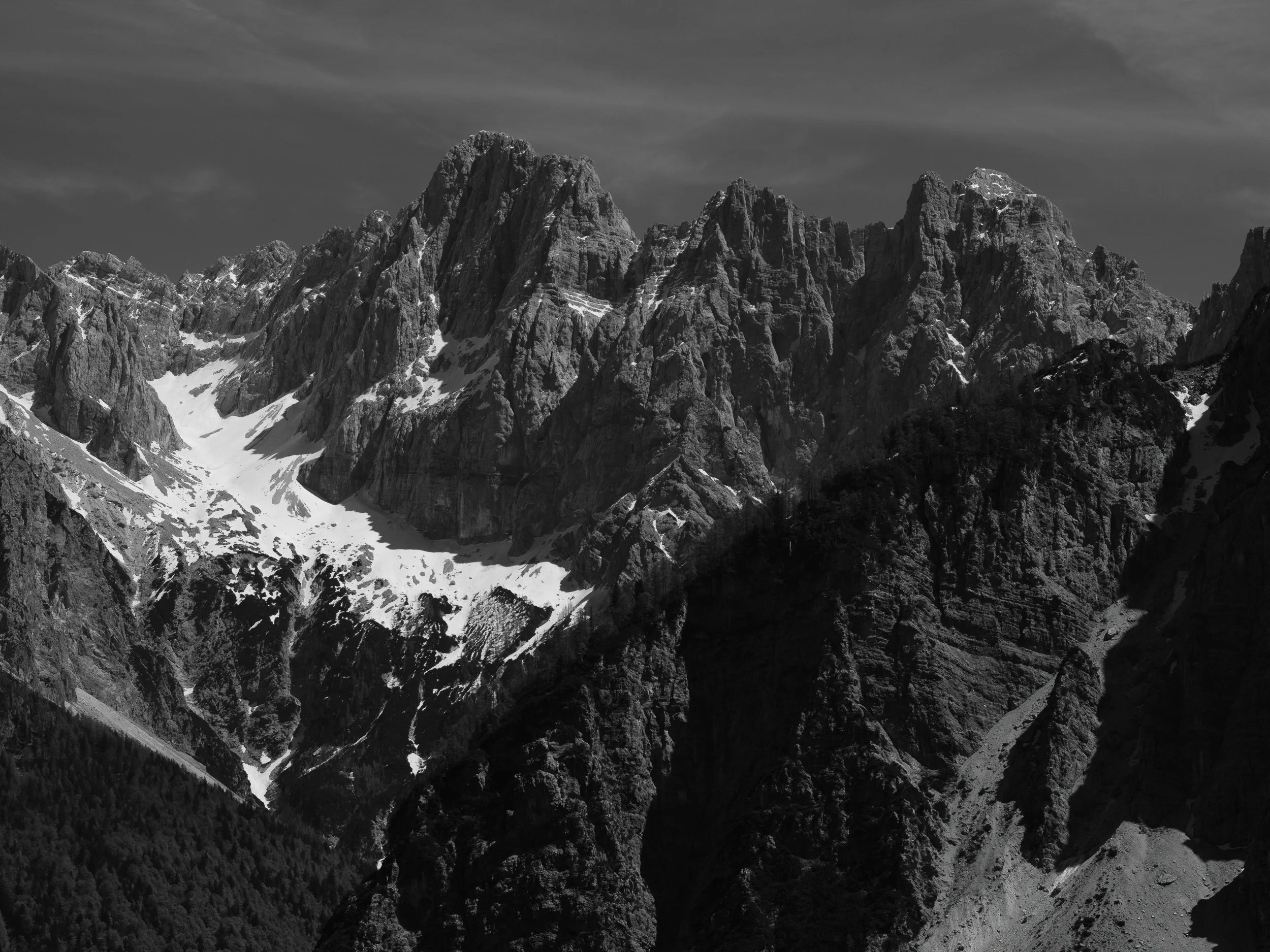 Berggipfel in einer Bergkette, mit Schneefeldern und Wolken am Himmel, in Schwarz-Weiß aufgenommen.