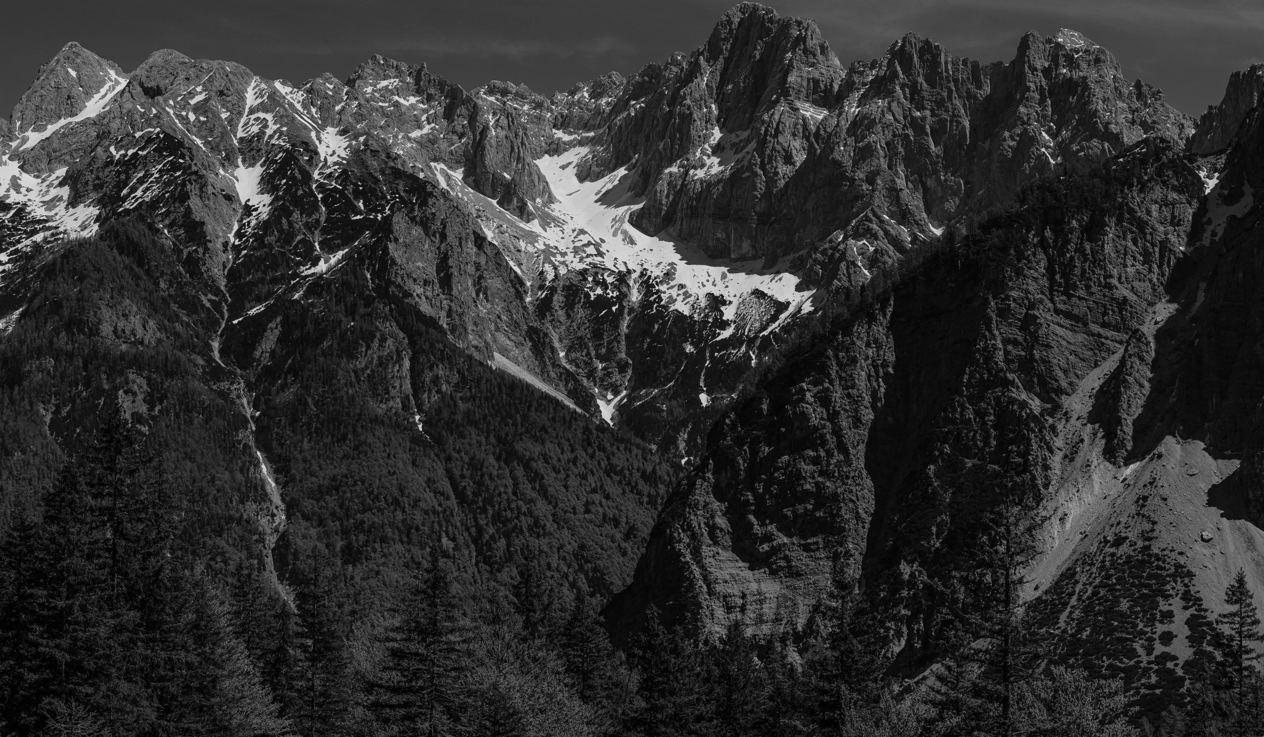 Schwarz-Weiß-Berglandschaft mit hohen, schroffen Gipfeln und schneebedeckten Flächen zwischen den Felsen, umgeben von Wäldern im unteren Bereich.