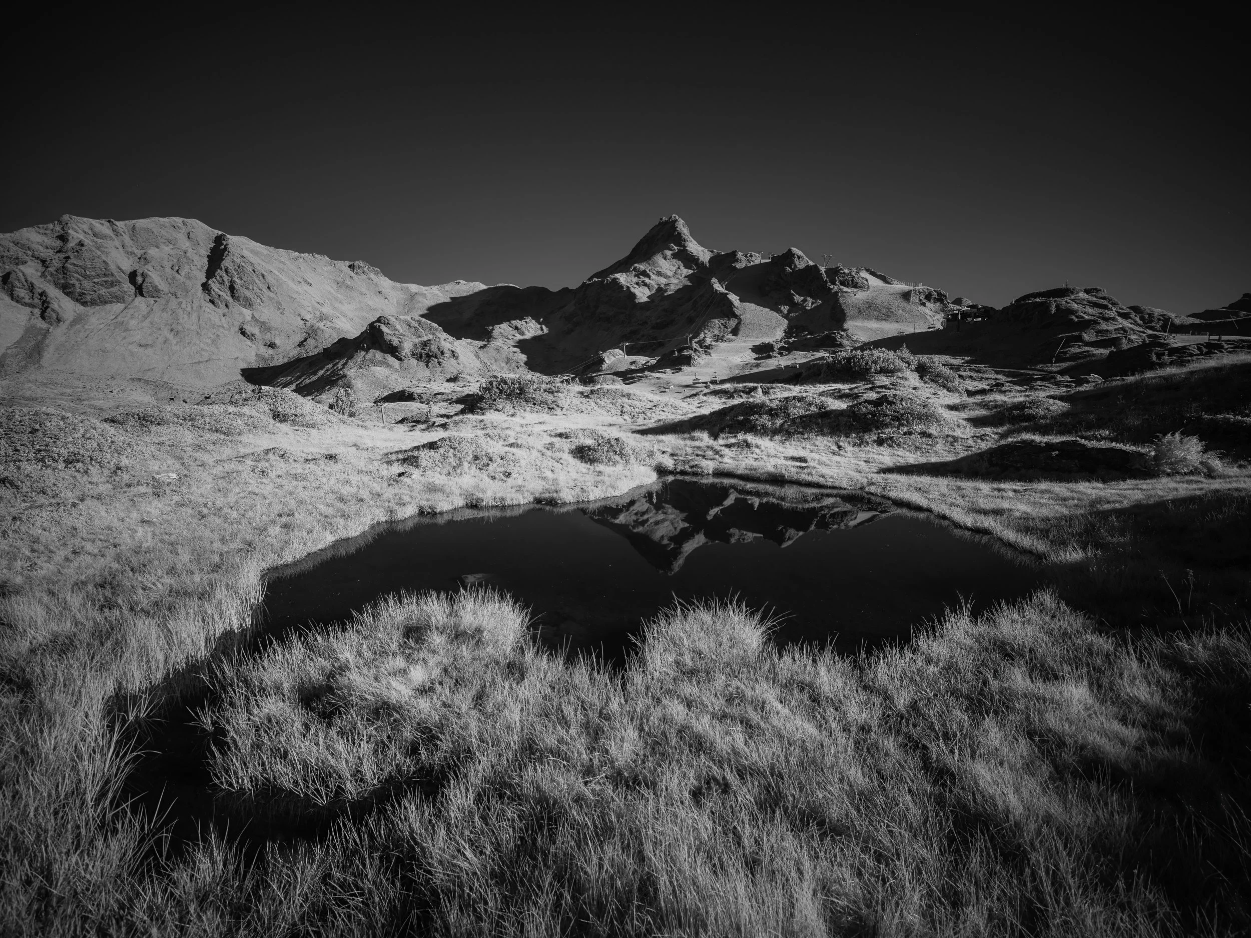Ein Bergpanorama mit einem See im Vordergrund, umgeben von Gras und felsigen Hügeln, in Schwarz-Weiß.