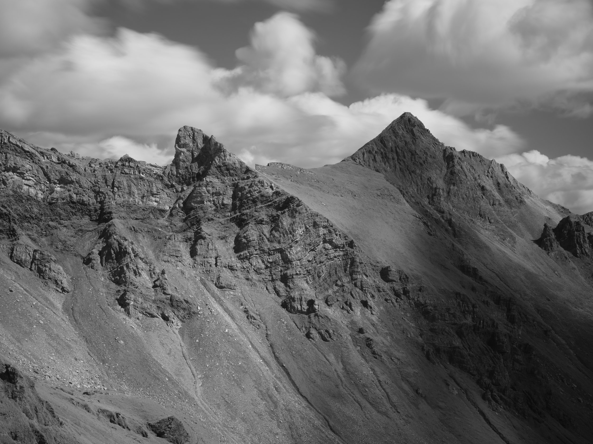 Schwarz-weiße Aufnahme von Bergen mit schroffen Gipfeln und Wolken am Himmel.