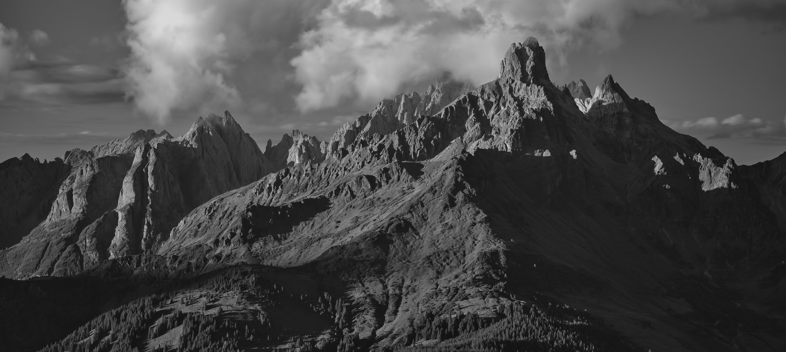 Schwarz-weiß-Bild von hohen Berggipfeln und Wolken am Himmel, mit einer Bergkette im Vordergrund.
