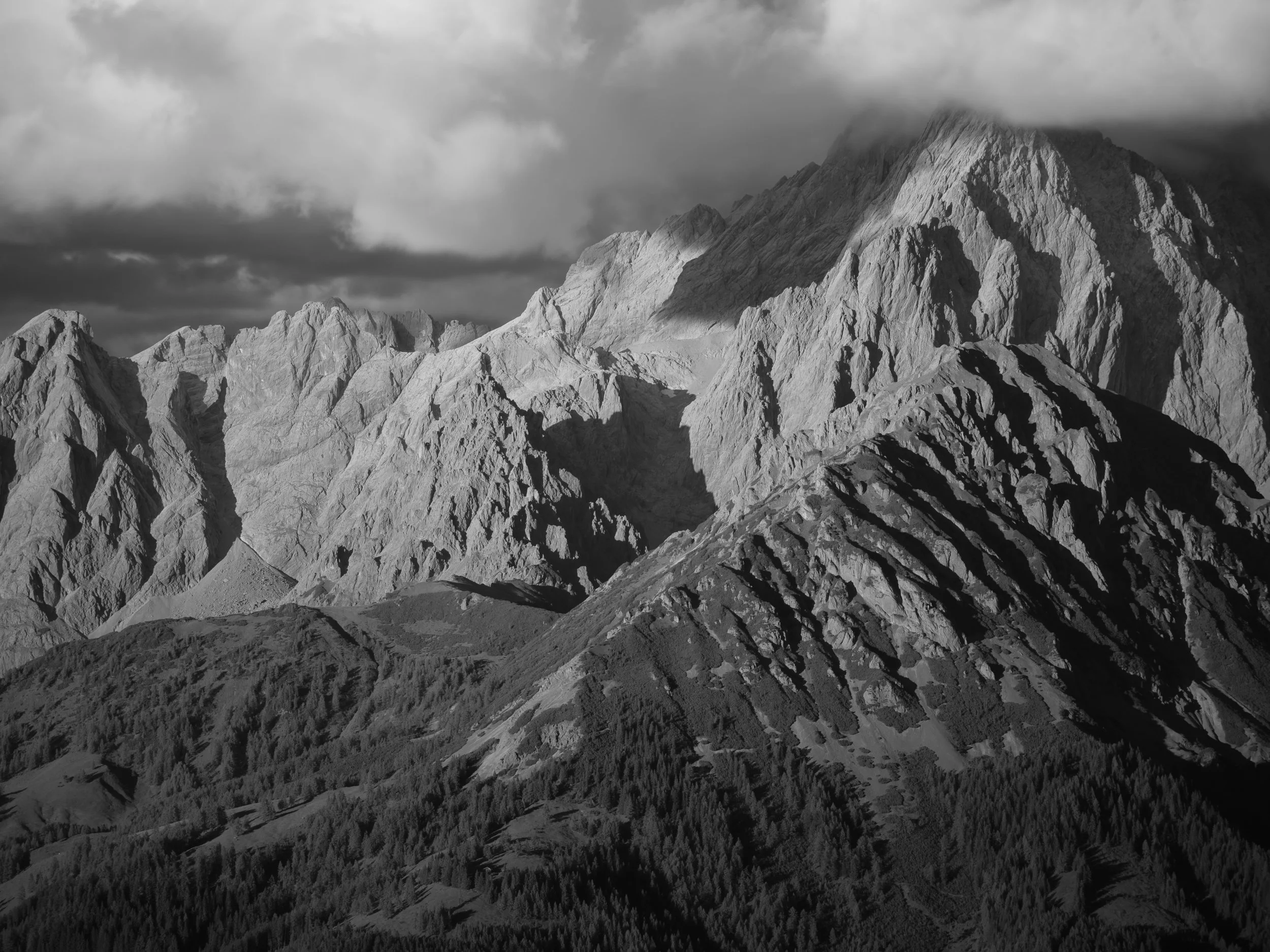 Bergkette mit schroffen, felsigen Gipfeln, teilweise bedeckt mit Schnee, unter dunklen Wolken.