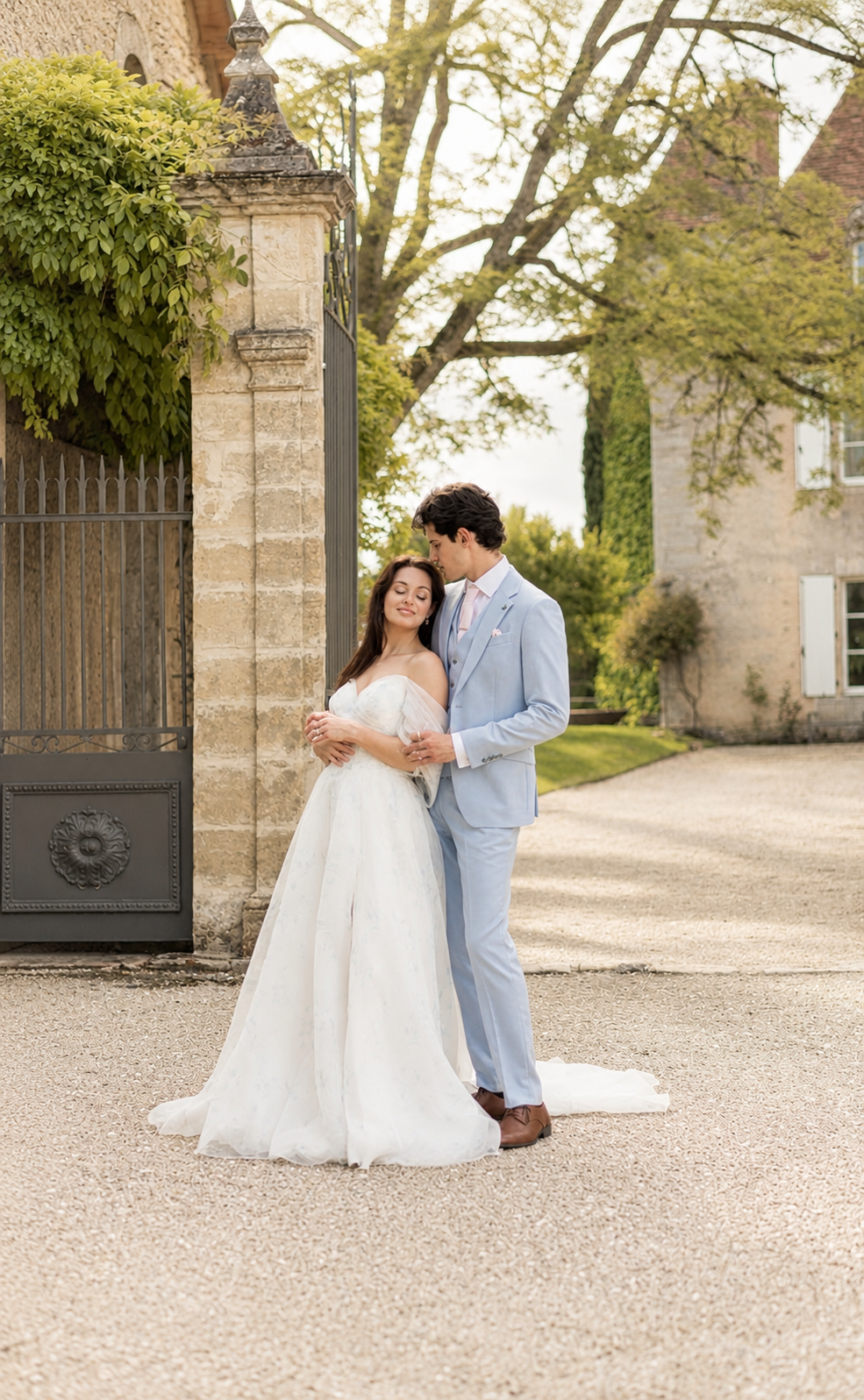A newlywed couple standing on a gravel driveway outside a stone building. The bride in a white wedding gown and the groom in a light blue suit share an intimate moment, with the groom gently kissing the bride's forehead. Greenery and trees surround t